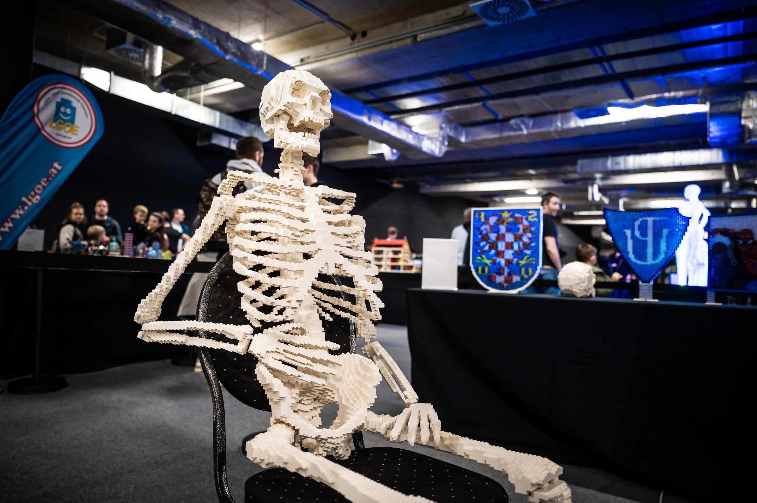 Eine Skulptur eines menschlichen Skeletts aus LEGO Steinen, sitzend auf einem schwarzen Stuhl, im Vordergrund. Im Hintergrund sind mehrere Personen und TDisplay-Elemente mit Logos und Wappen bei einer Veranstaltung zu sehen.