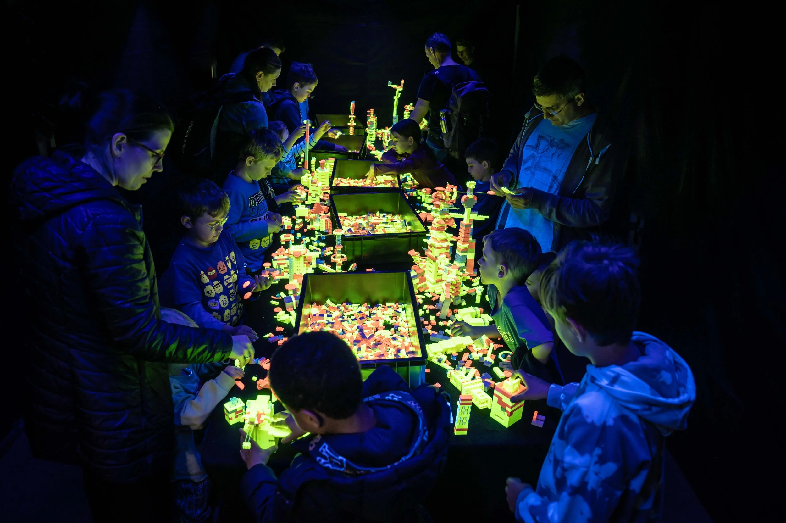 Kinder und Erwachsene spielen mit leuchtenden Lego-Steinen in einer dunklen Umgebung.