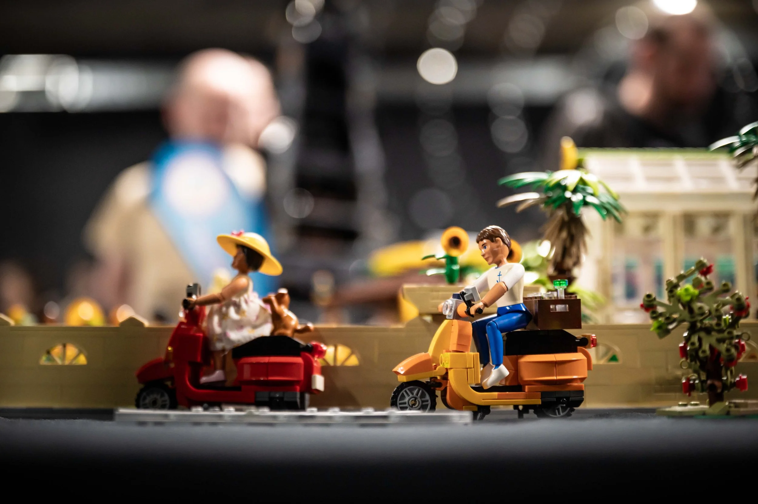 Eine Lego-Szene mit Figuren auf einem Wagen, umgeben von Pflanzen, im Hintergrund sind verschwommene Menschen. Es handelt sich um eine kreative Miniatur-Darstellung.