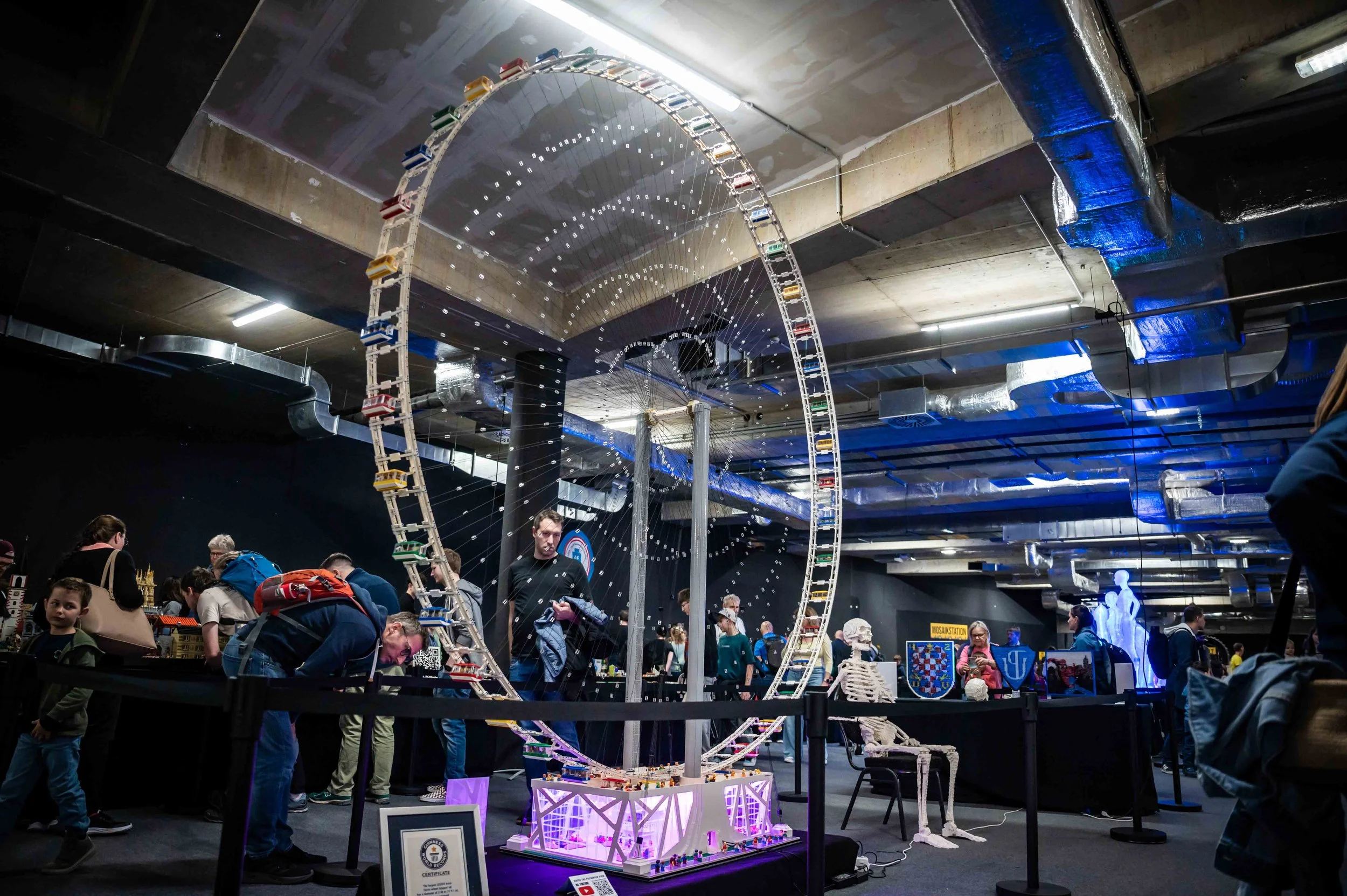 Innenraum eines Erlebnisparks mit großer, leuchtender Achterbahn aus LEGO, umgeben von Menschen und Ausstellungsstücken, darunter Skelette und Wappen.