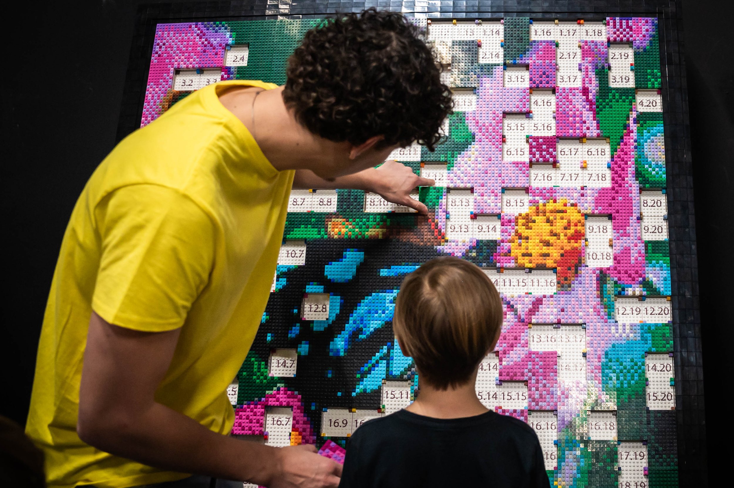 Menschen, darunter zwei Kinder, betrachten ein großes LEGO-Bild. Ein Mann in gelbem T-Shirt zeigt auf das LEGO-Bild, während die Kinder es ansehen.