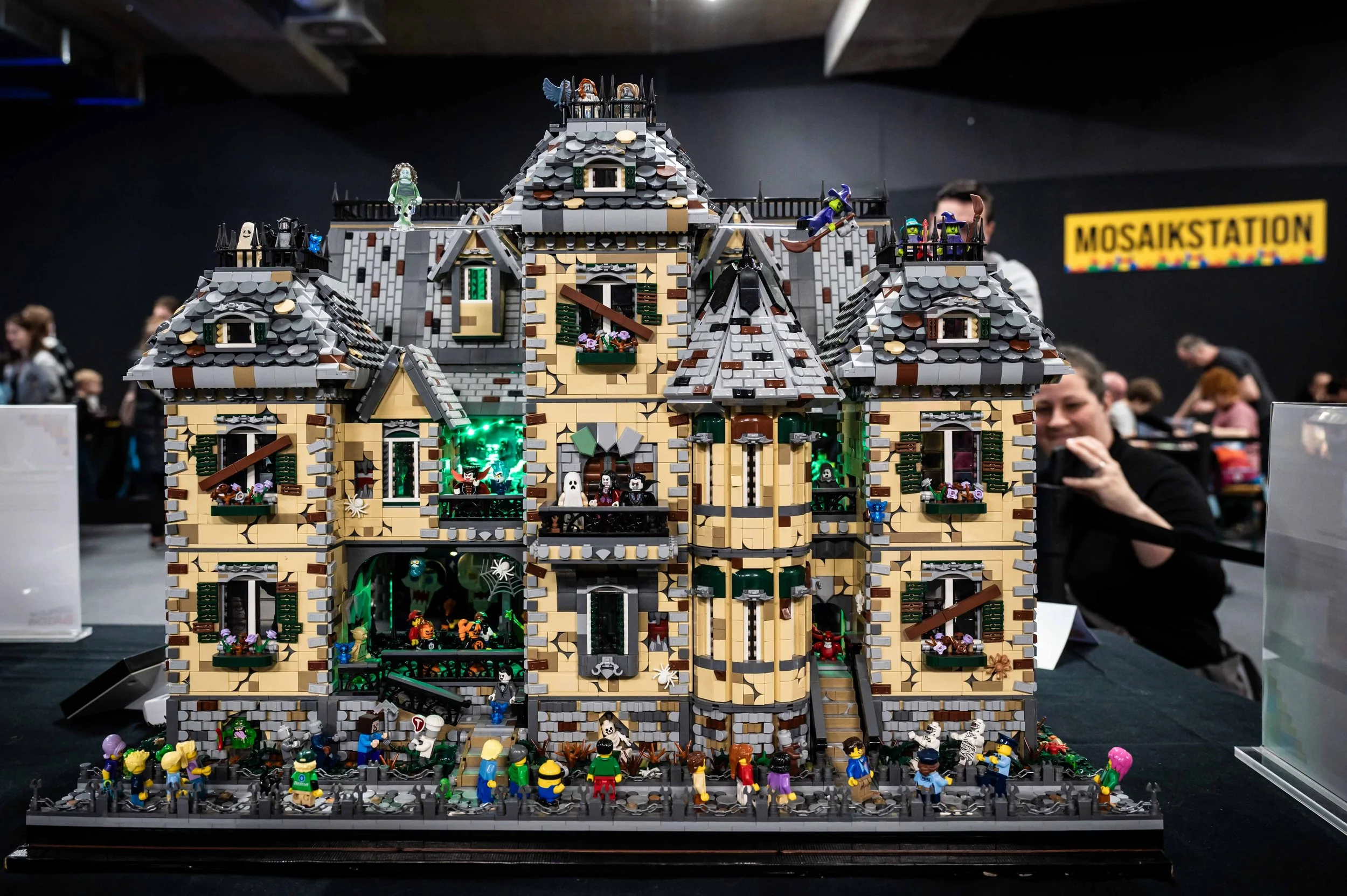 Eine detailreiche Halloween-Burg aus LEGO mit Hexen, Gespenstern, Zombies und anderen Figuren, geschmückt mit Spinnenweben und Skeletten, auf einer Ausstellung mit mehreren Menschen im Hintergrund.