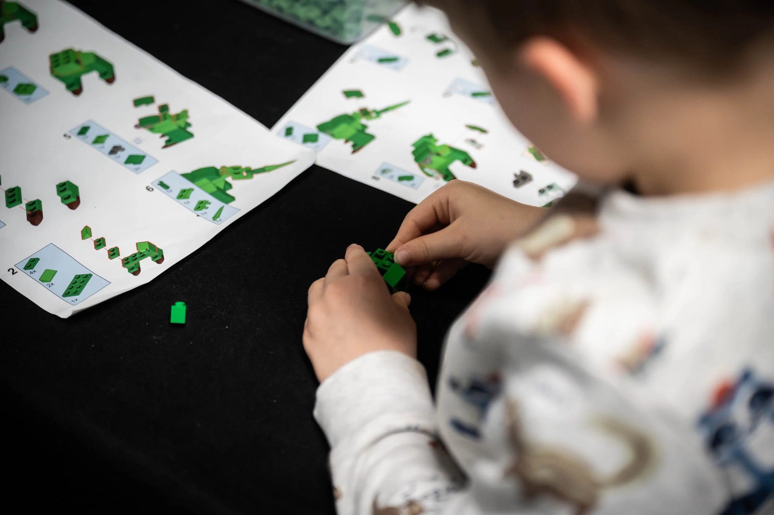 Ein Kind baut mit grünen LEGO-Steinen auf einem schwarzen Tisch während der Anleitung, die auf Papier vor ihm liegt.