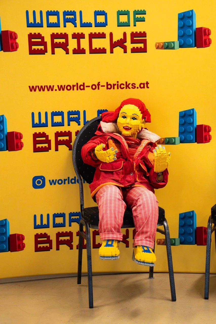 Eine Figur, die komplett aus LEGO-Steinen besteht, sitzt auf einem Stuhl vor einem gelben Hintergrund mit dem Schriftzug 'World of Bricks' und einer Webseite. Die LEGO-Figur trägt eine rote Jacke, rote und weiße Hose, und gelbe LEGO-Füße und Hände, m