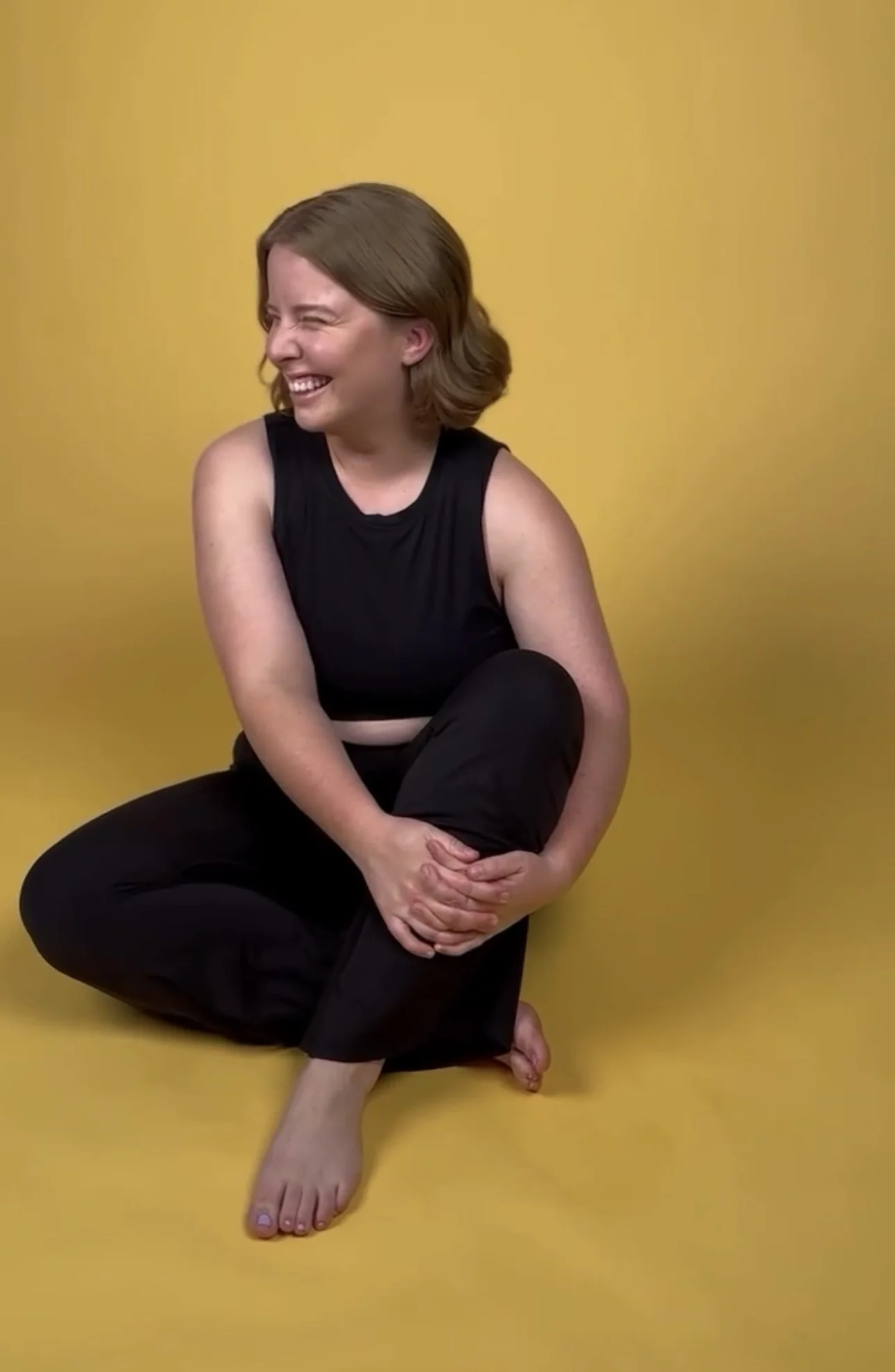 A woman with short brown hair, wearing a black sleeveless top and black pants, sitting on the floor against a yellow background. She is laughing and holding her ankle while crossing her legs.