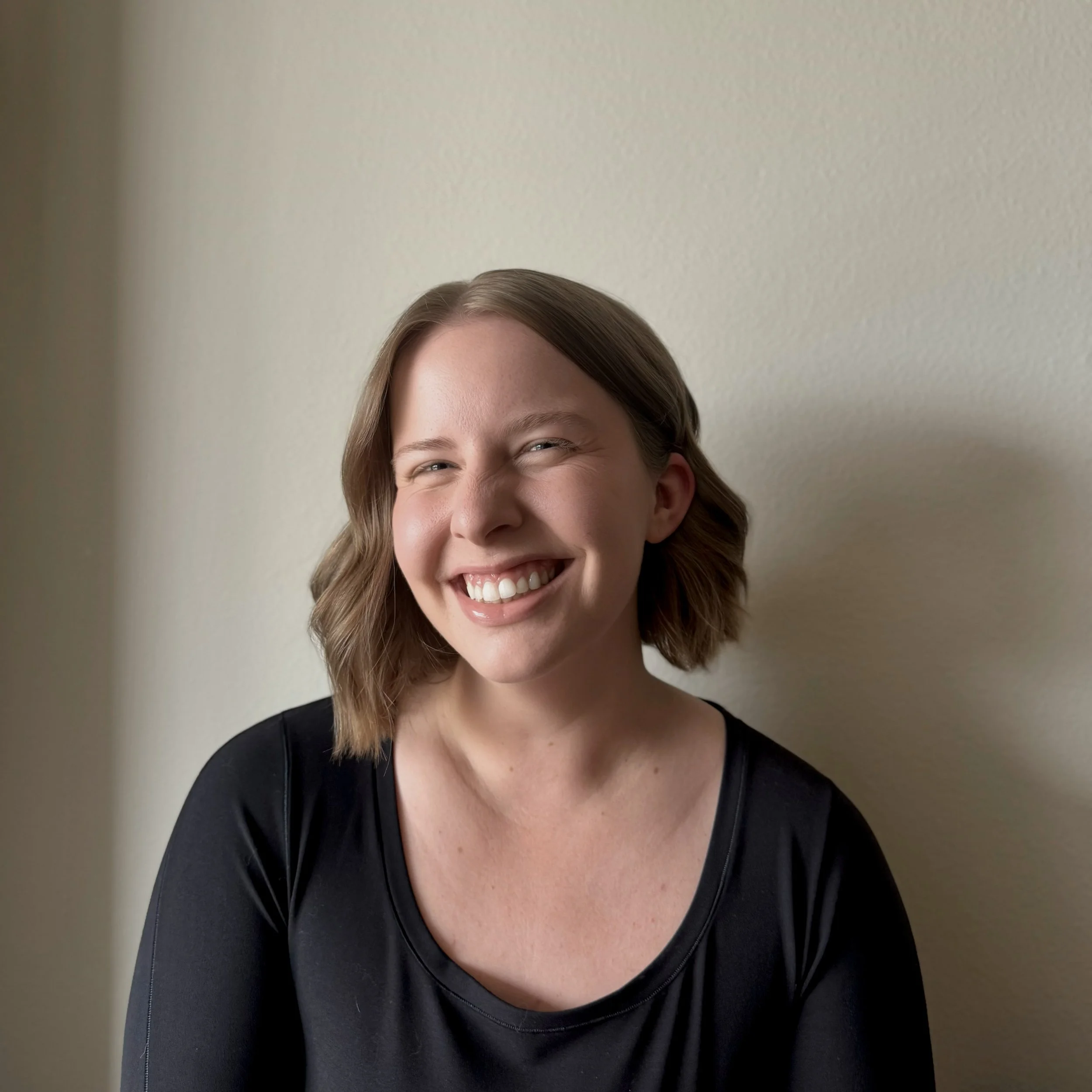 A woman with shoulder-length light brown hair, smiling and wearing a black top, standing against a beige wall.