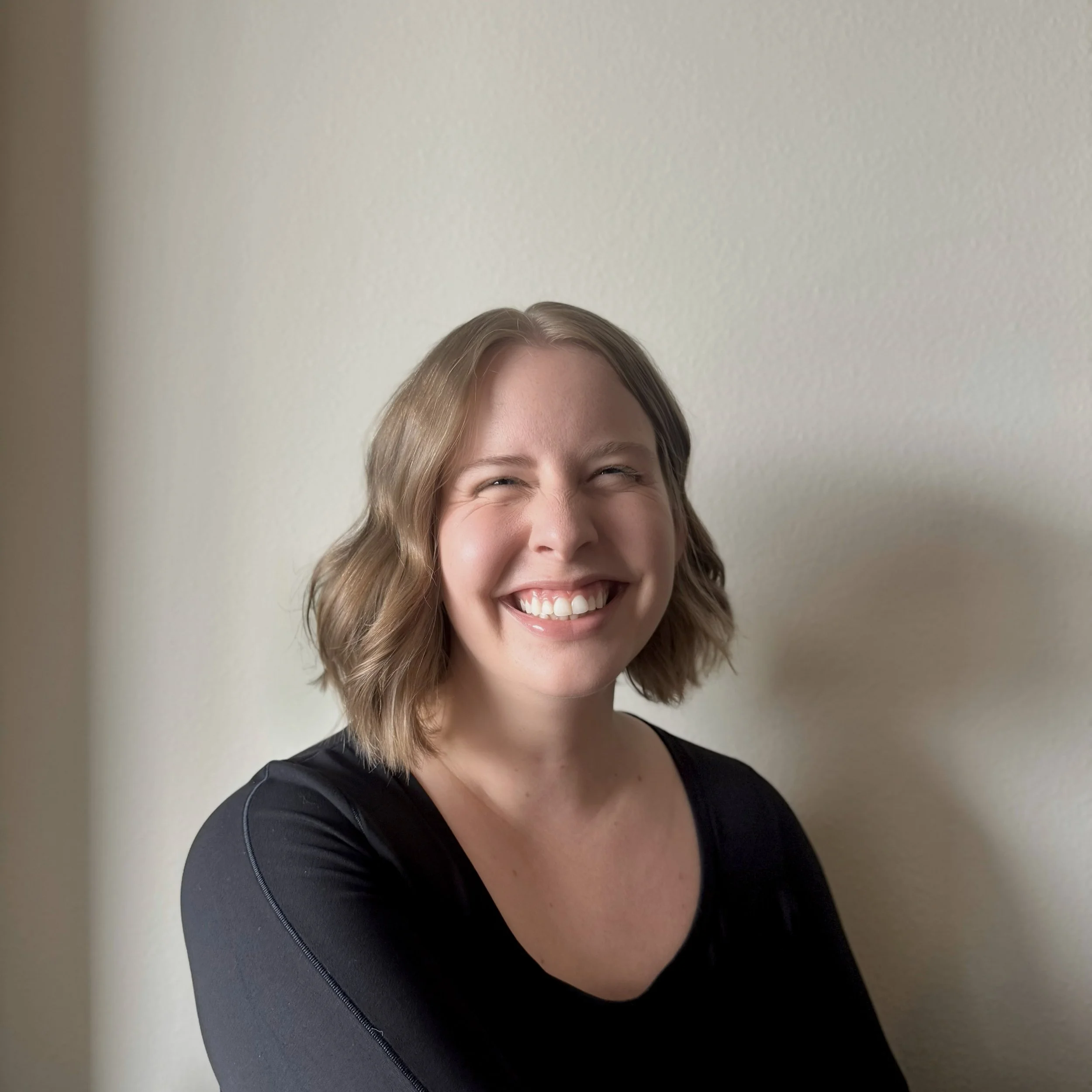 Amelia Sweet, a smiling woman with shoulder-length wavy brown hair, wearing a black top, standing against a plain light-colored wall.