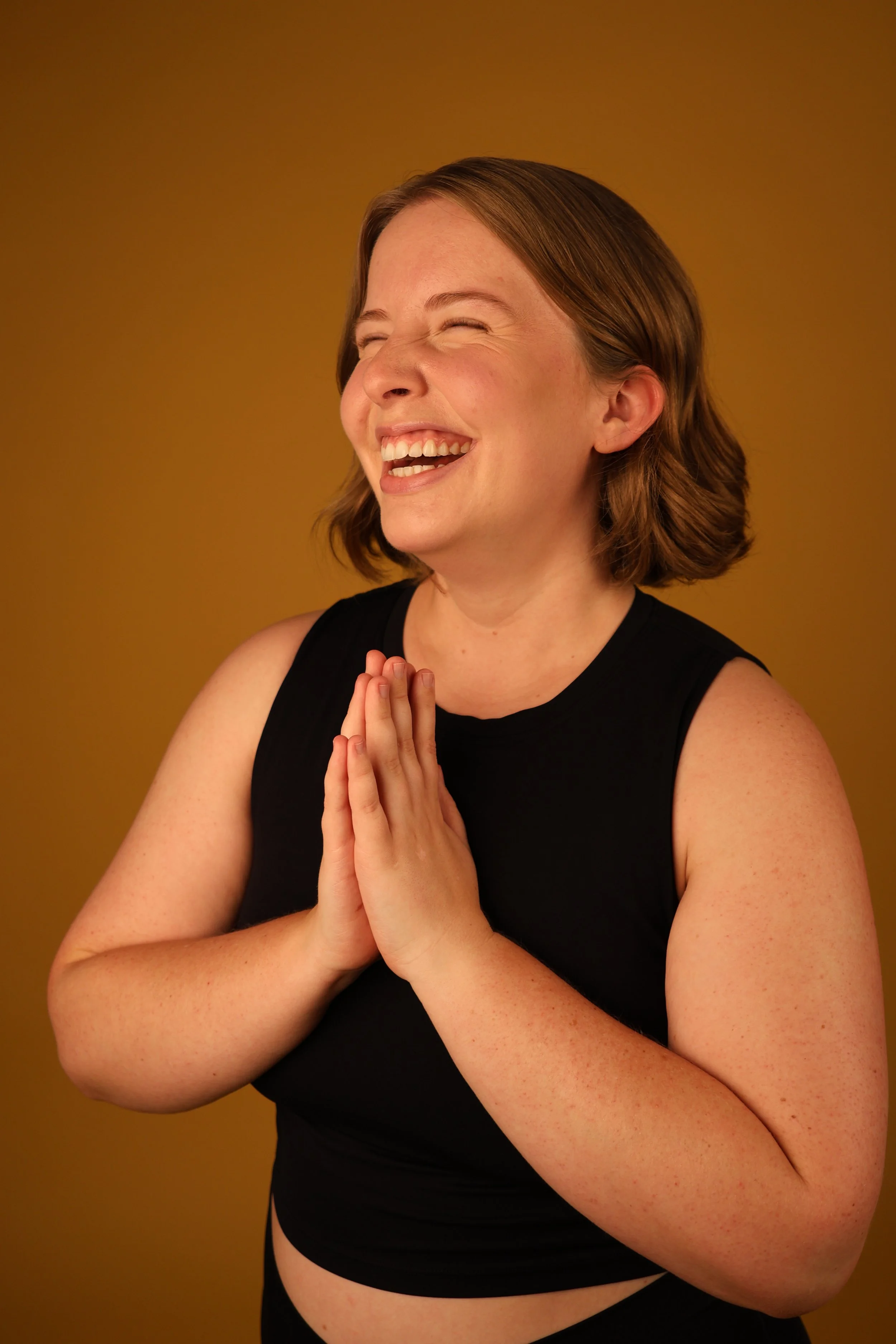 Amelia Sweet - A woman with short brown hair laughing with eyes closed, hands pressed together in a prayer or gratitude pose, wearing a black sleeveless top, against a brown background.
