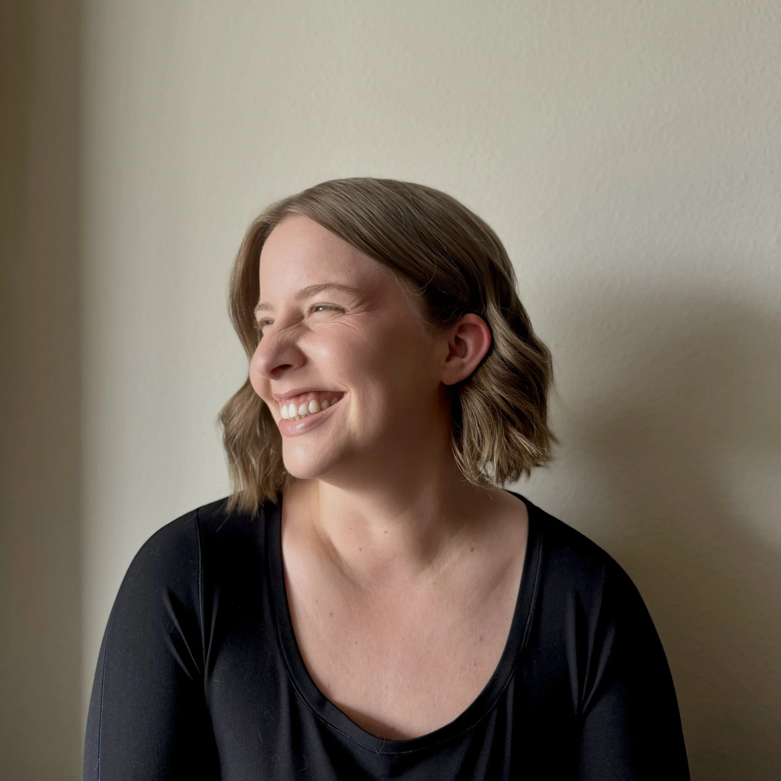 A woman with shoulder-length brown hair smiling and looking to her left against a plain light-colored wall.