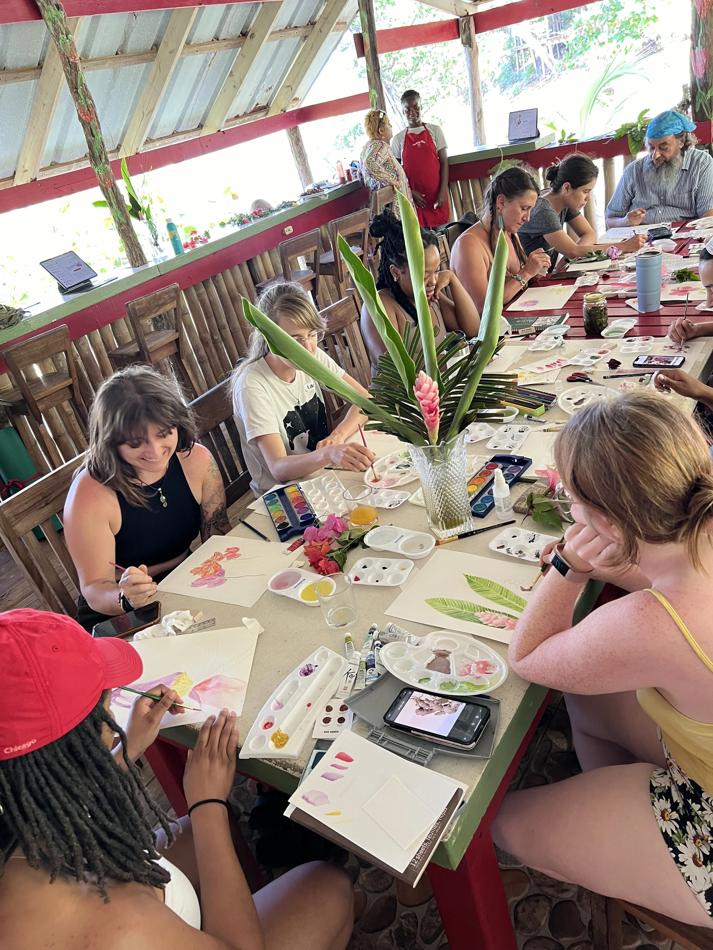 Group of people participating in a watercolor painting class in a rustic, open-air setting with natural light, surrounded by potted plants and a large tropical flower arrangement.