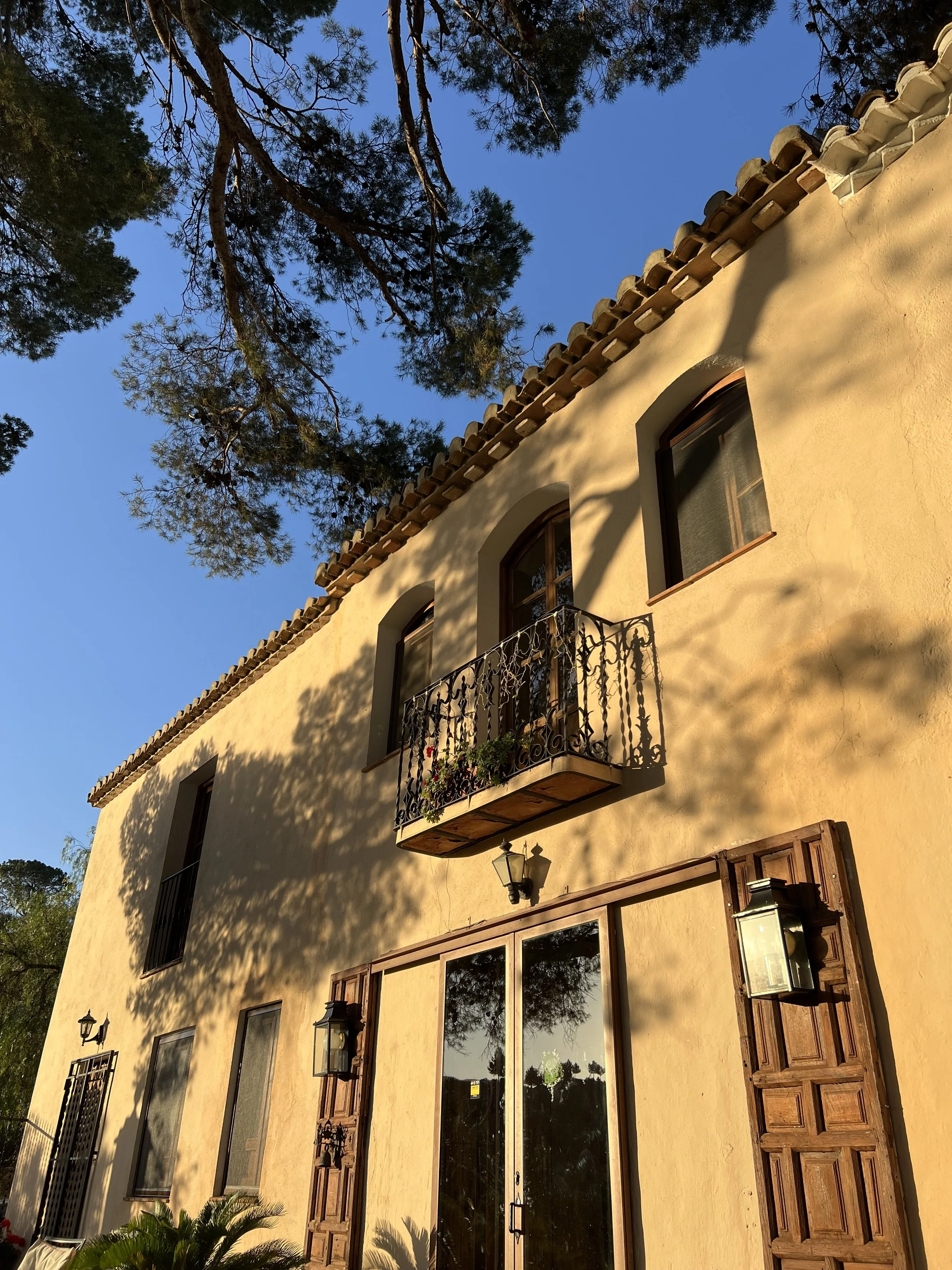 A two-story beige stucco house with wooden shutters, a small balcony with wrought iron railing, and multiple outdoor wall-mounted lanterns, with tree branches casting shadows on its facade during daylight.