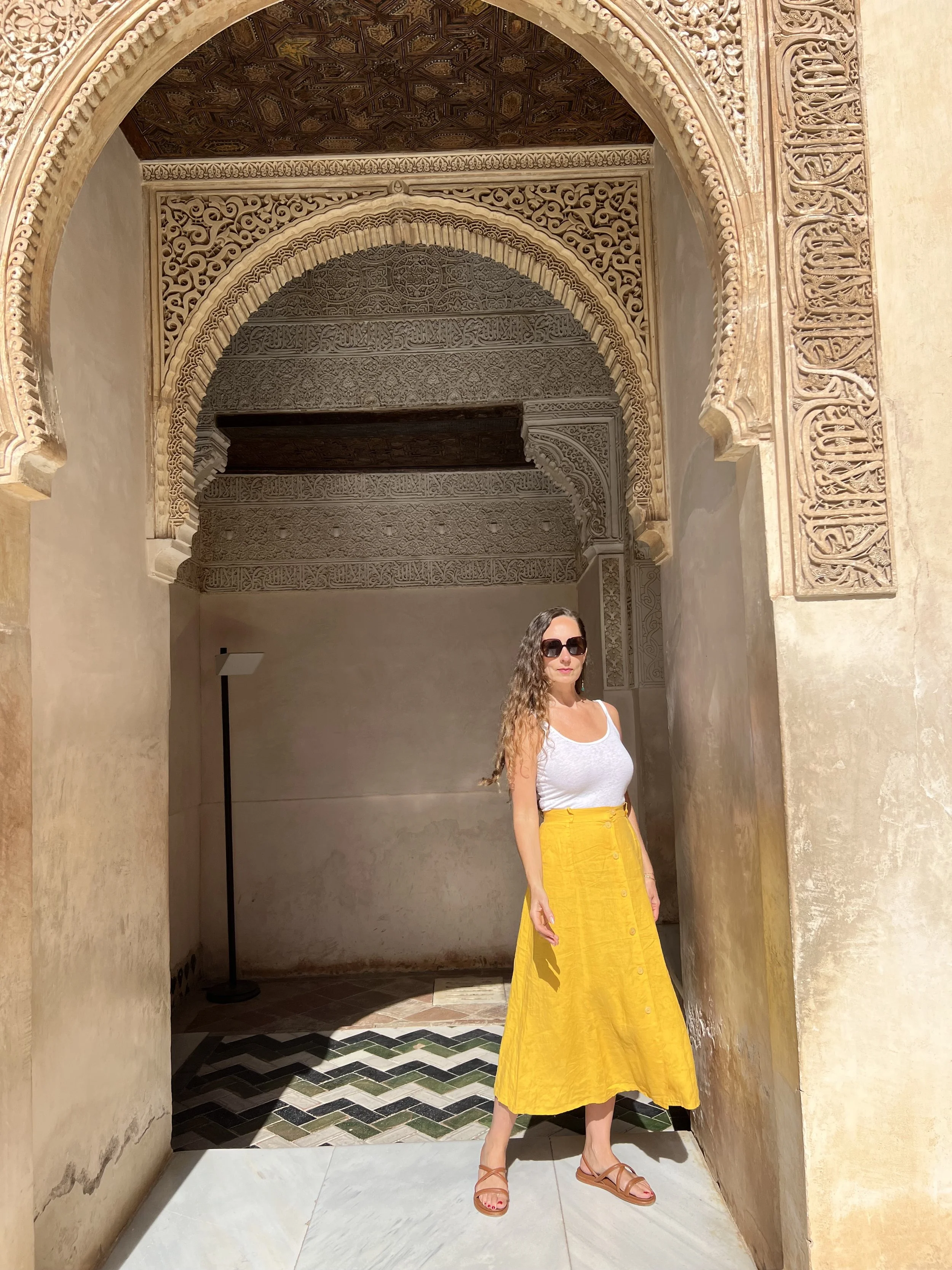 A woman in sunglasses, a white tank top, a yellow skirt, and sandals standing in a doorway with intricate Moroccan-style archway and ceiling carvings.
