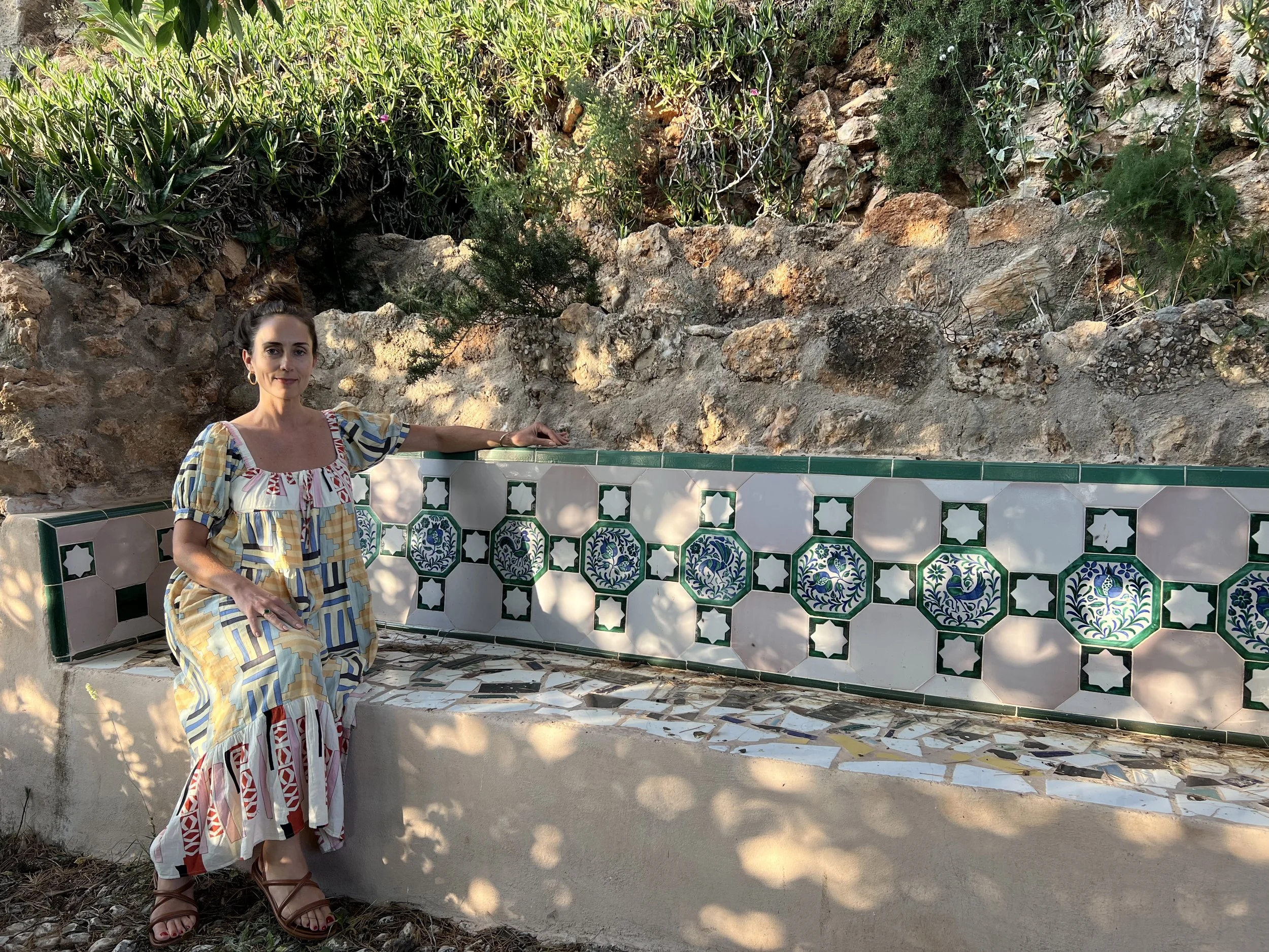 A woman sitting on a decorative tiled bench outdoors, with a rocky and green background.
