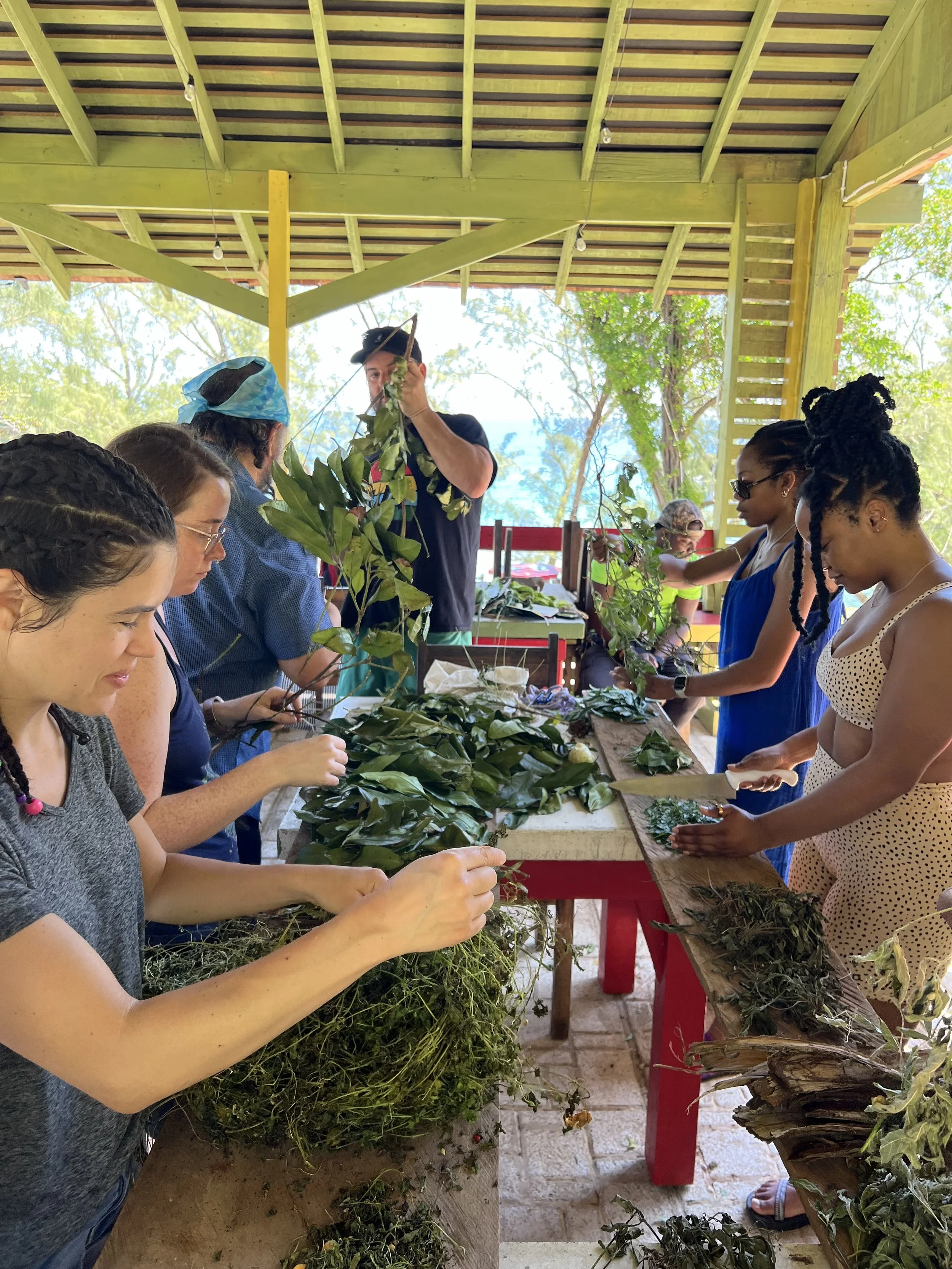 Group of people working together on a craft project involving green leaves and plant materials at a covered outdoor table