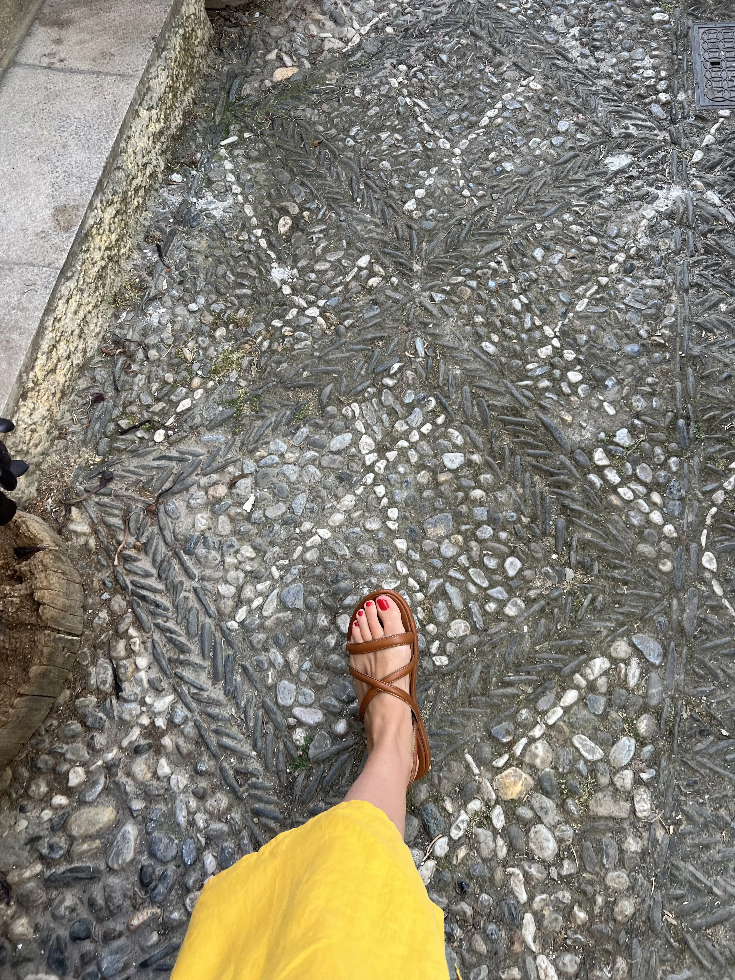A person's foot wearing a brown sandal with red toenail polish, visible on a yellow skirt, walking on decorative stone pavement with leaf and pebble patterns.