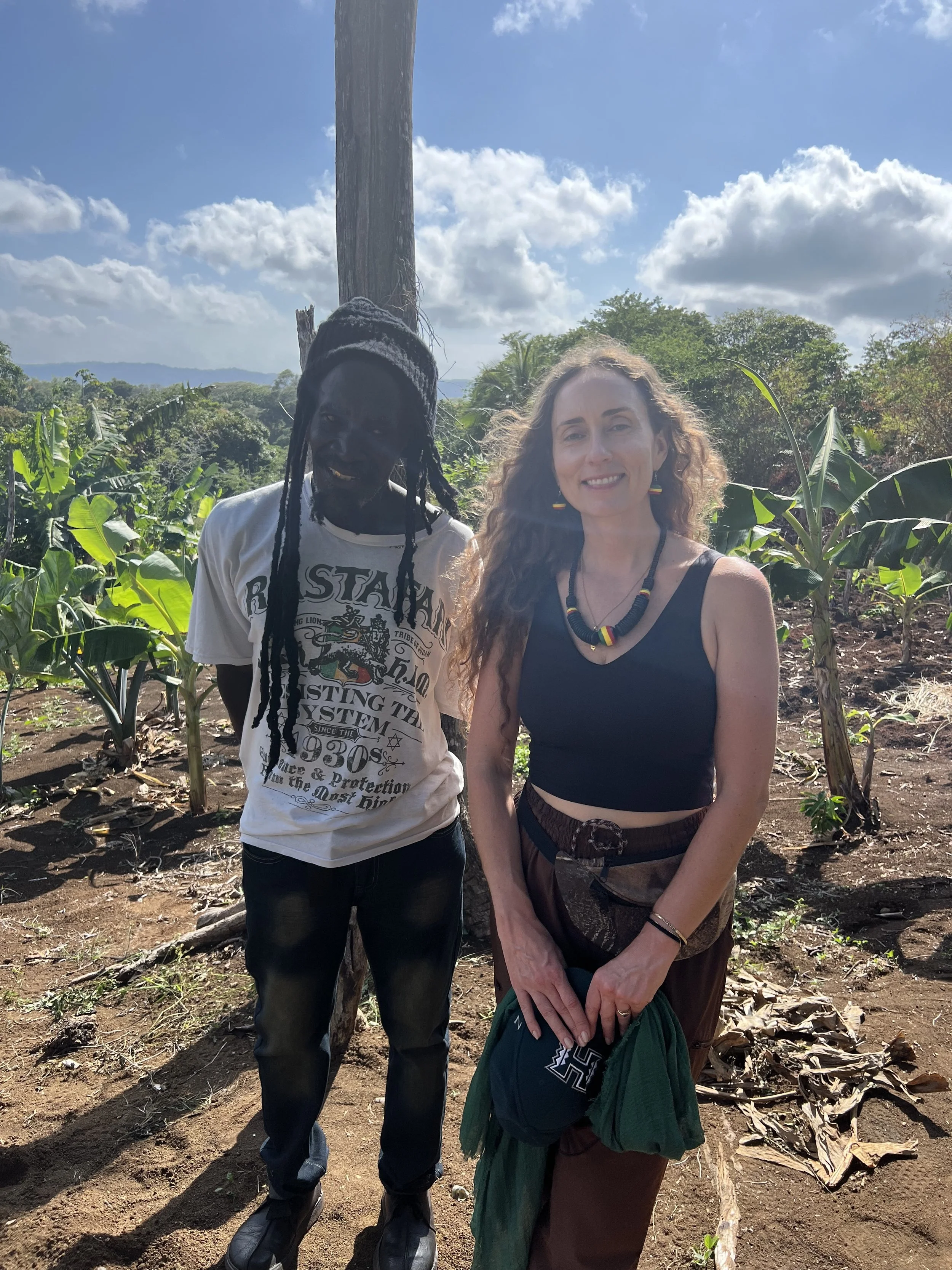 Two women standing in a lush green field of banana plants, smiling at the camera, with blue skies and scattered clouds overhead.