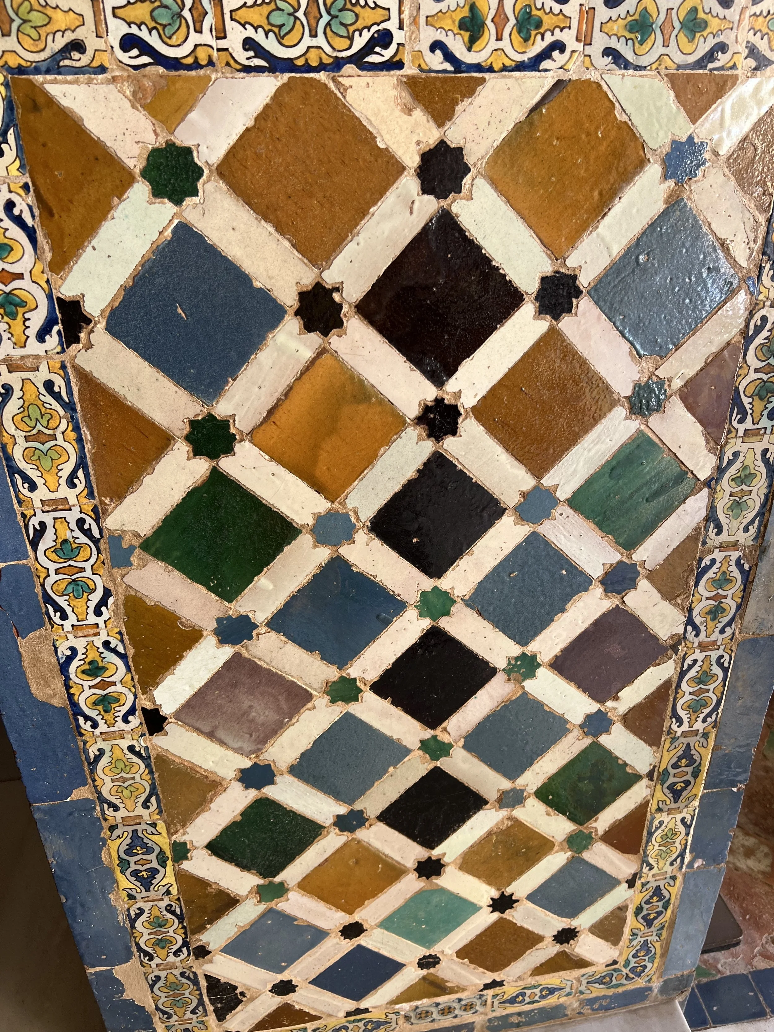 Close-up of a decorative mosaic tile ceiling with geometric patterns, colorful tiles including blue, green, yellow, black, and white, surrounded by intricate floral border designs.