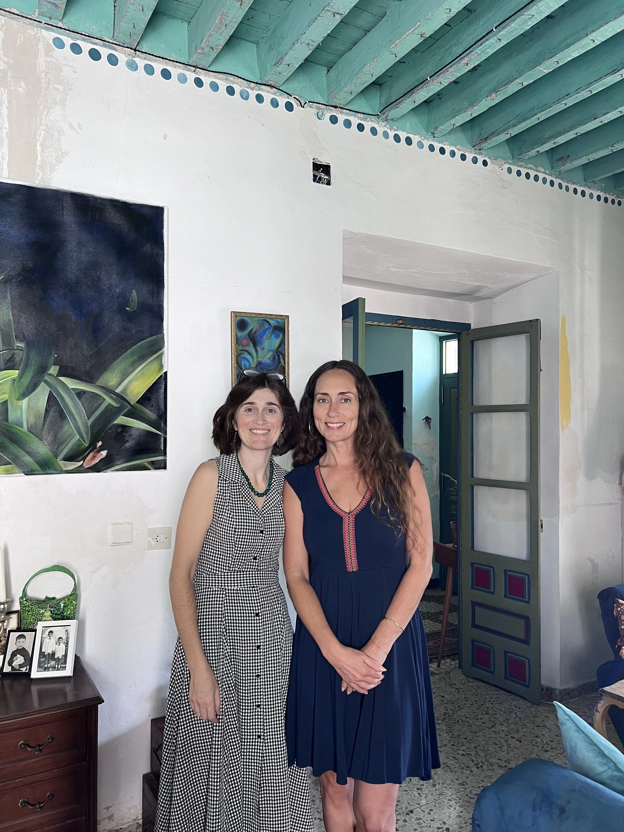 Two women standing inside a house, smiling at the camera. One is wearing a black and white checkered dress, and the other is wearing a navy blue dress with pink accents. The room has white walls, teal ceiling beams, and artwork on the wall.