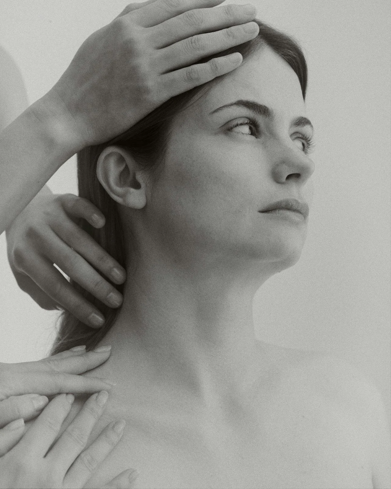 A black and white photo of a woman with a serene expression, her head slightly tilted, and multiple hands gently touching her face and neck.