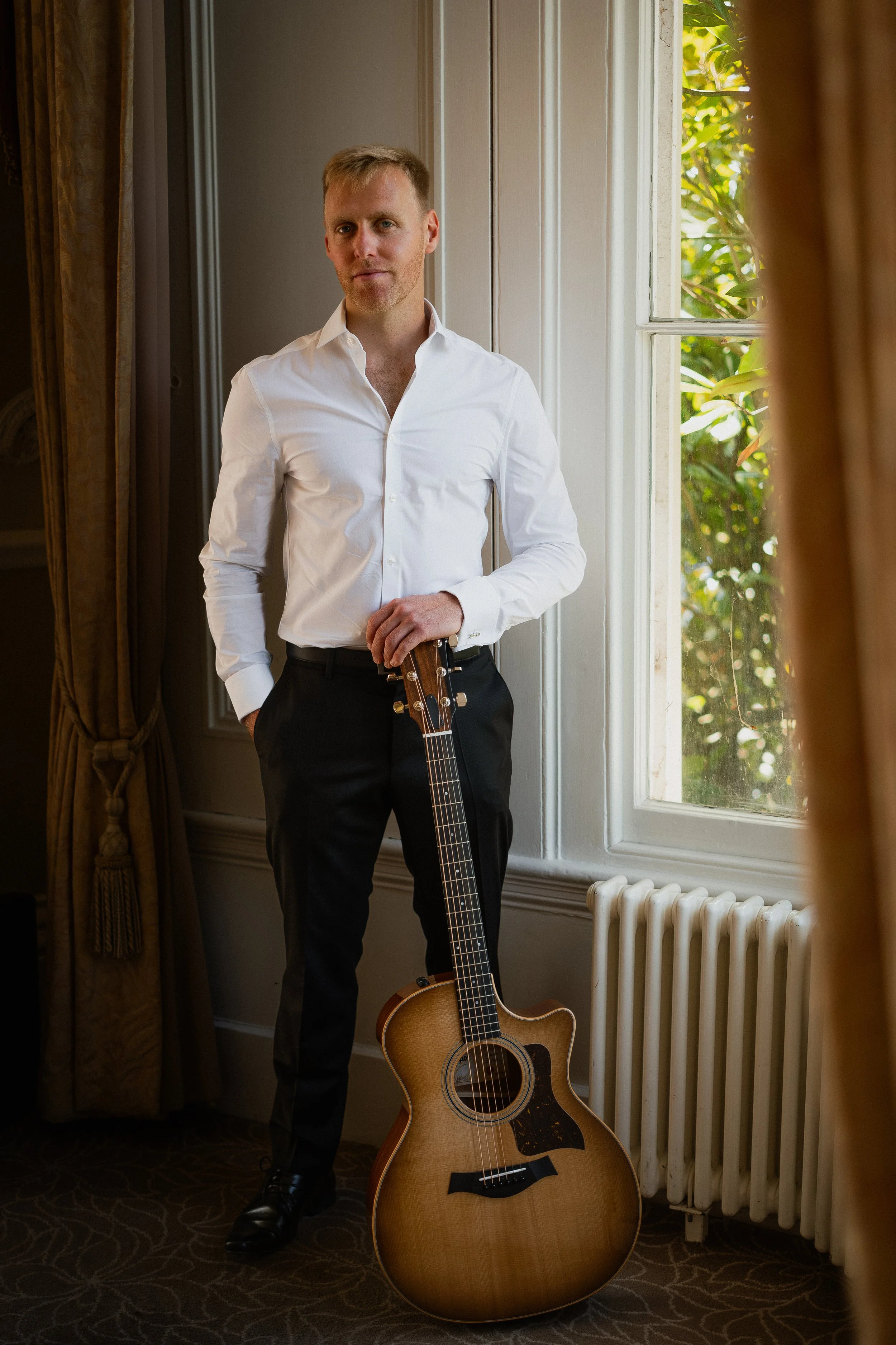 A man in a white dress shirt and black pants standing indoors near a window, holding an acoustic guitar.