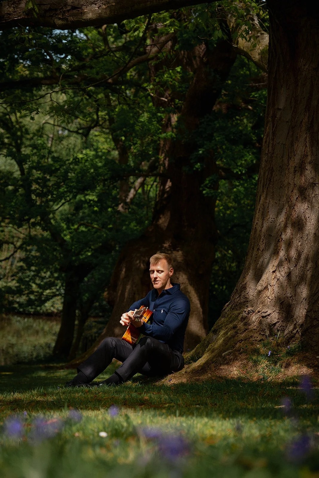 A man relaxes under a tree, playing an acoustic guitar, creating a tranquil atmosphere in the natural surroundings.