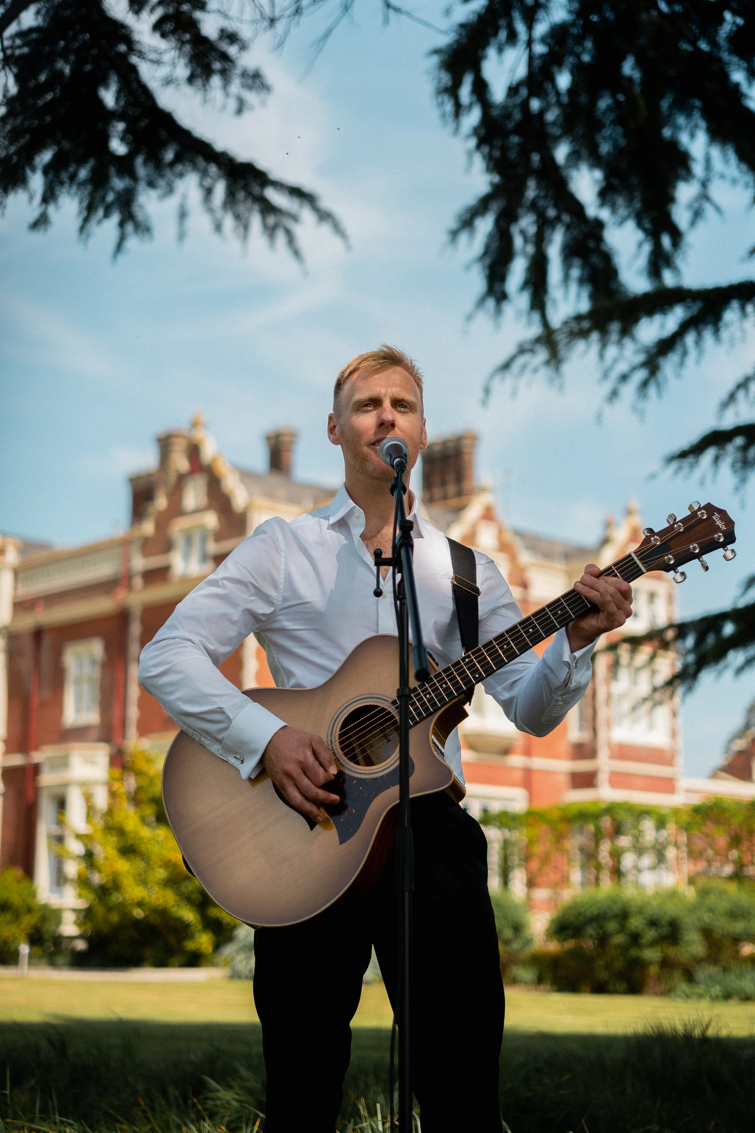 A man in a white shirt playing an acoustic guitar and singing into a microphone outdoors with a large historic brick building in the background.