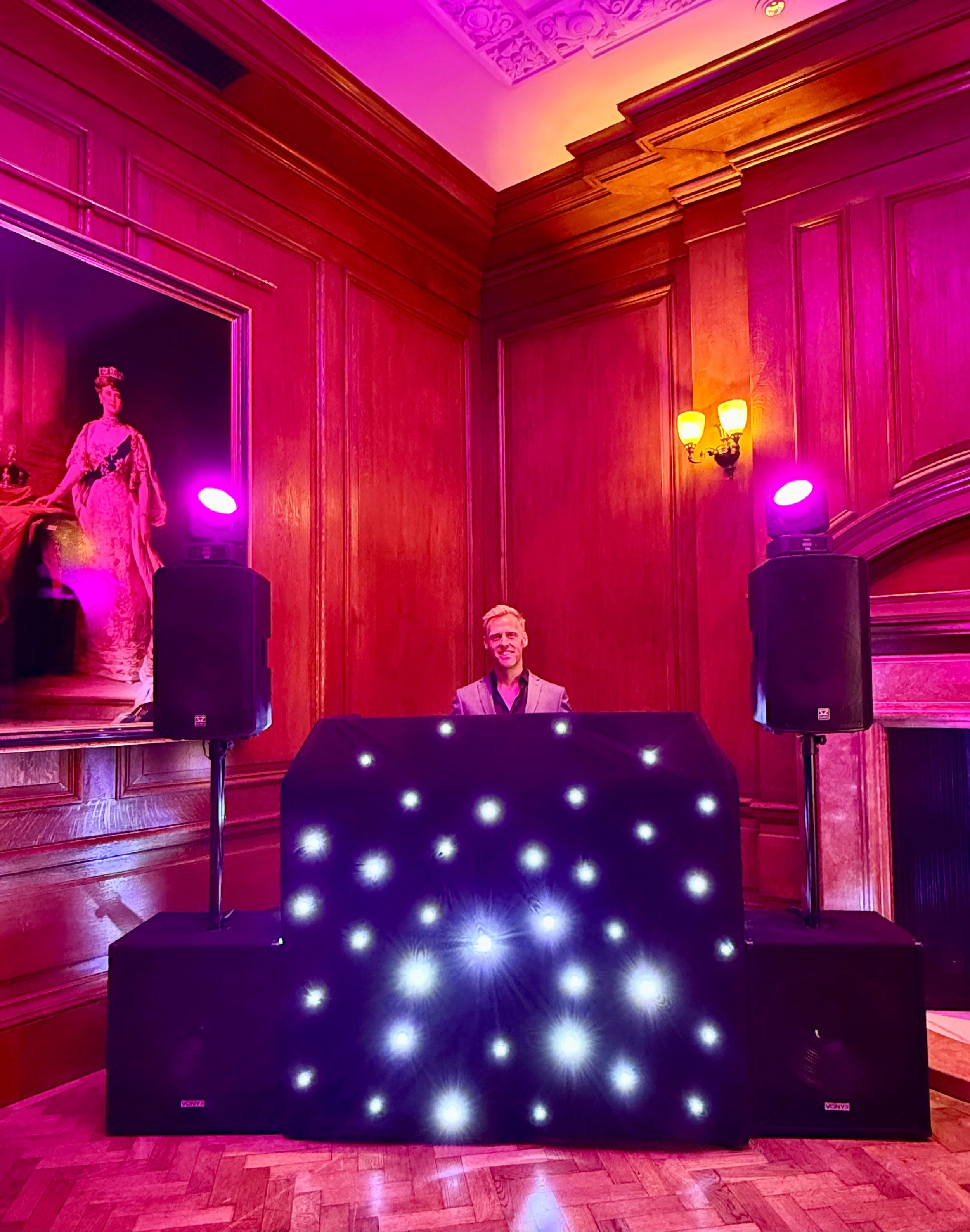 A man standing behind a DJ booth decorated with white lights, with a wooden panel wall and a painting of a woman in a dress on the wall, illuminated by pink and purple stage lighting.