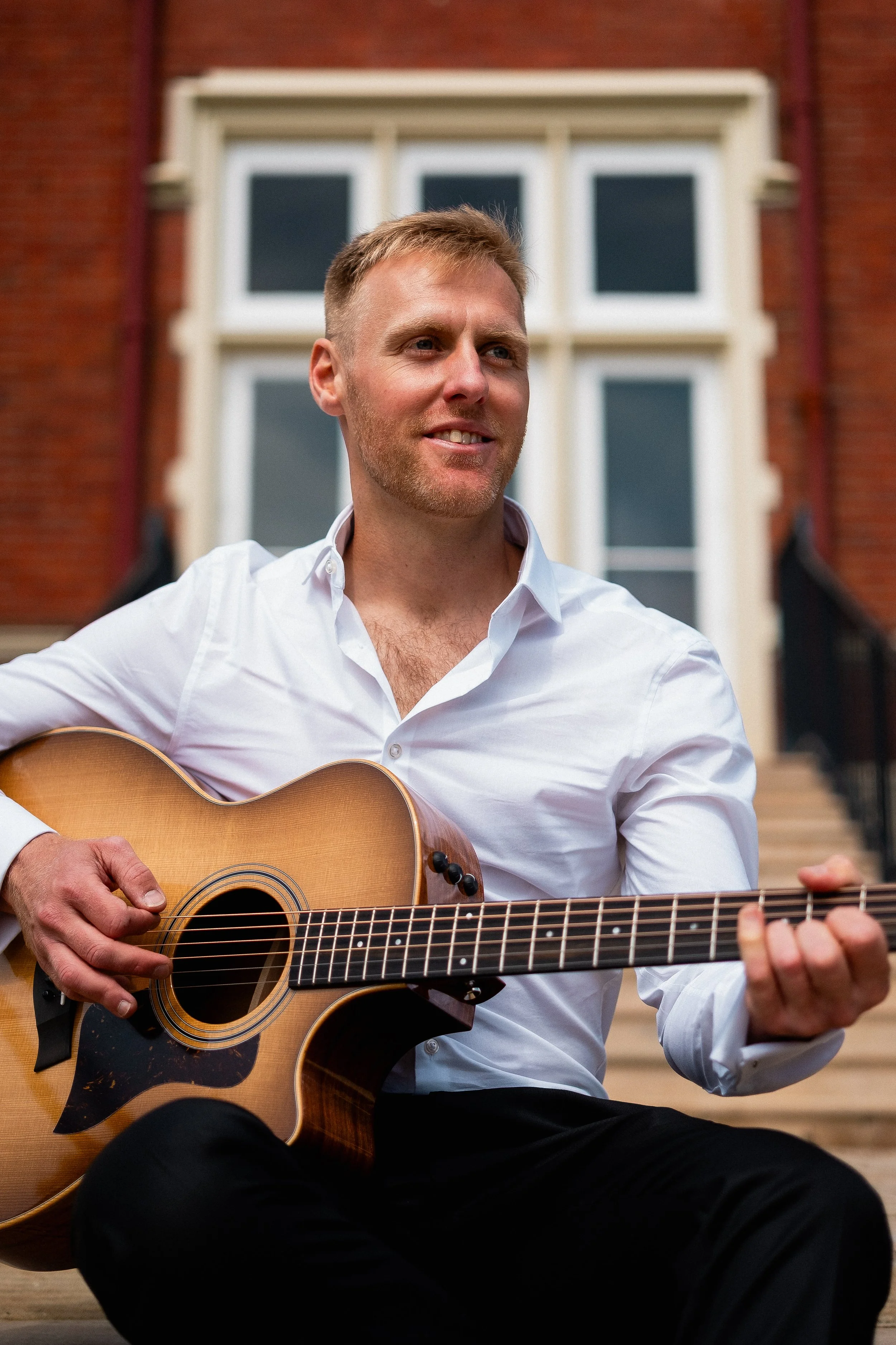 A musician sits on the steps strumming guitar at a luxury wedding venue while smiling to his audience.