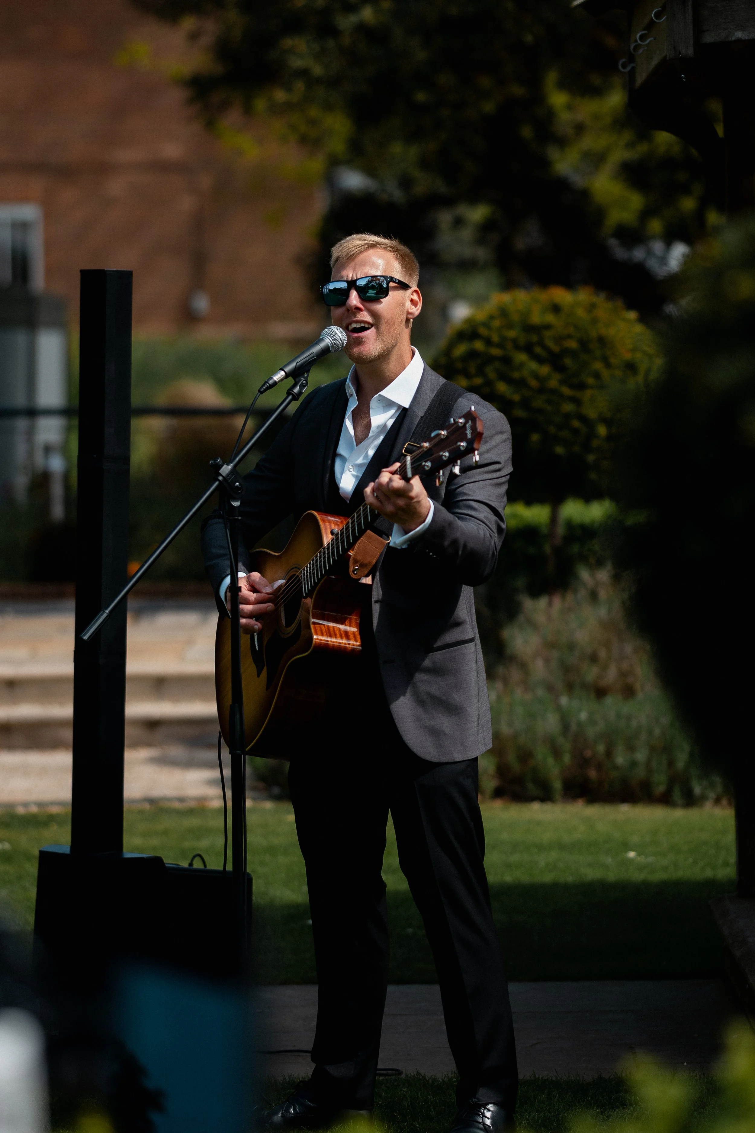 Man performing outdoors, playing guitar and singing into microphone, wearing sunglasses and a suit, with greenery and a brick building in the background.