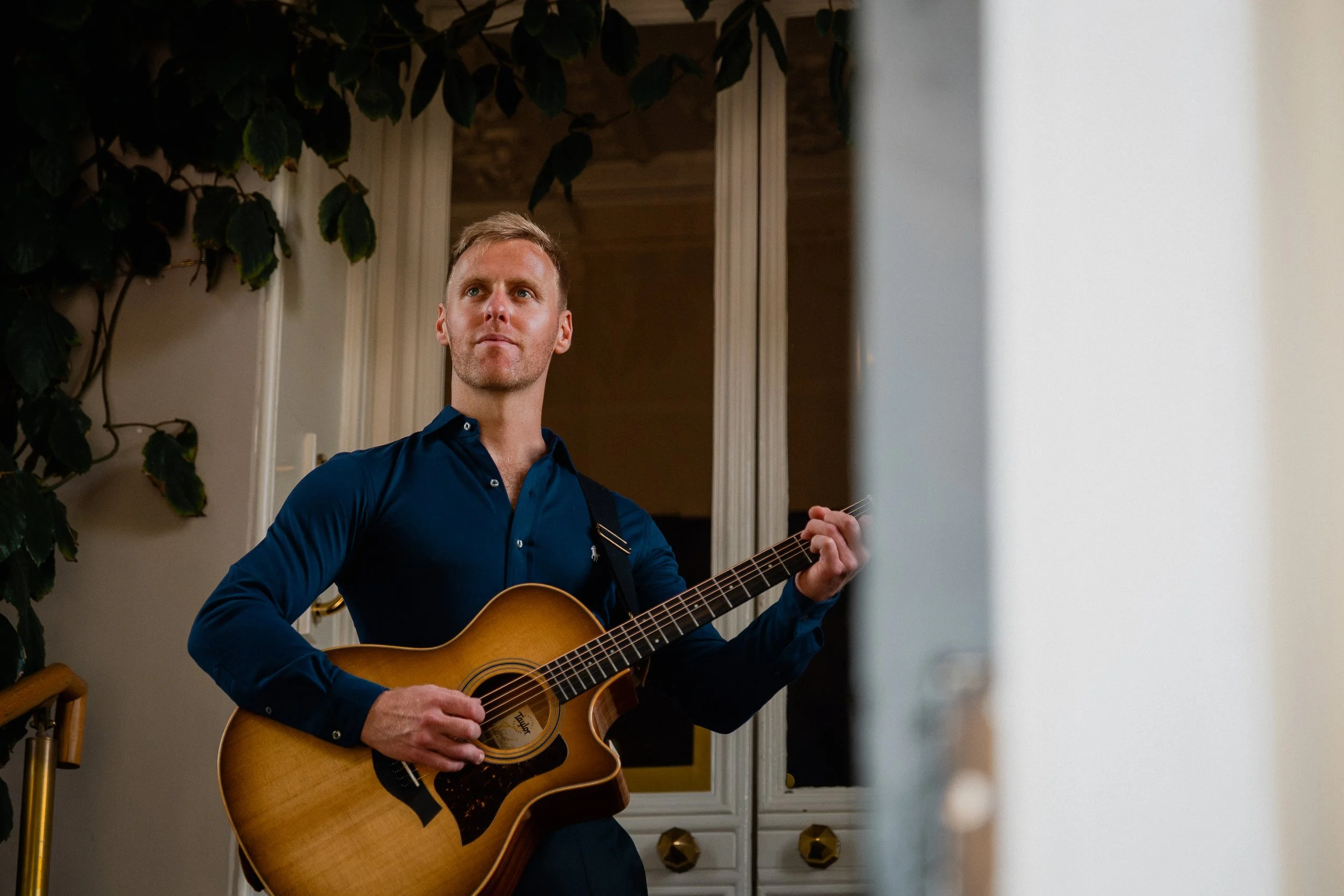 A man in a blue shirt playing an acoustic guitar indoors, standing in front of a white door with glass panels, surrounded by plants.