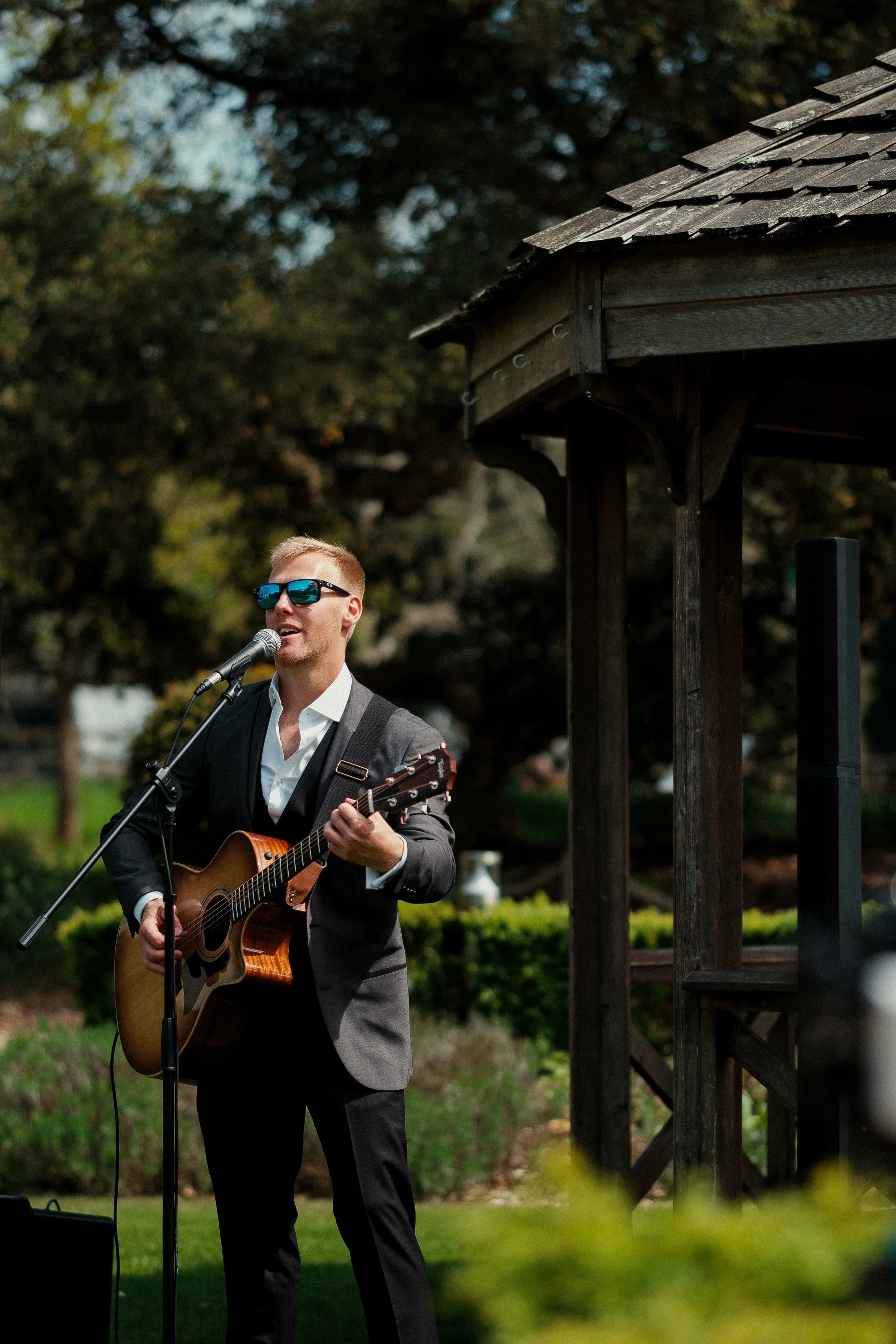 A man wearing sunglasses and a suit is playing an acoustic guitar and singing into a microphone outdoors.
