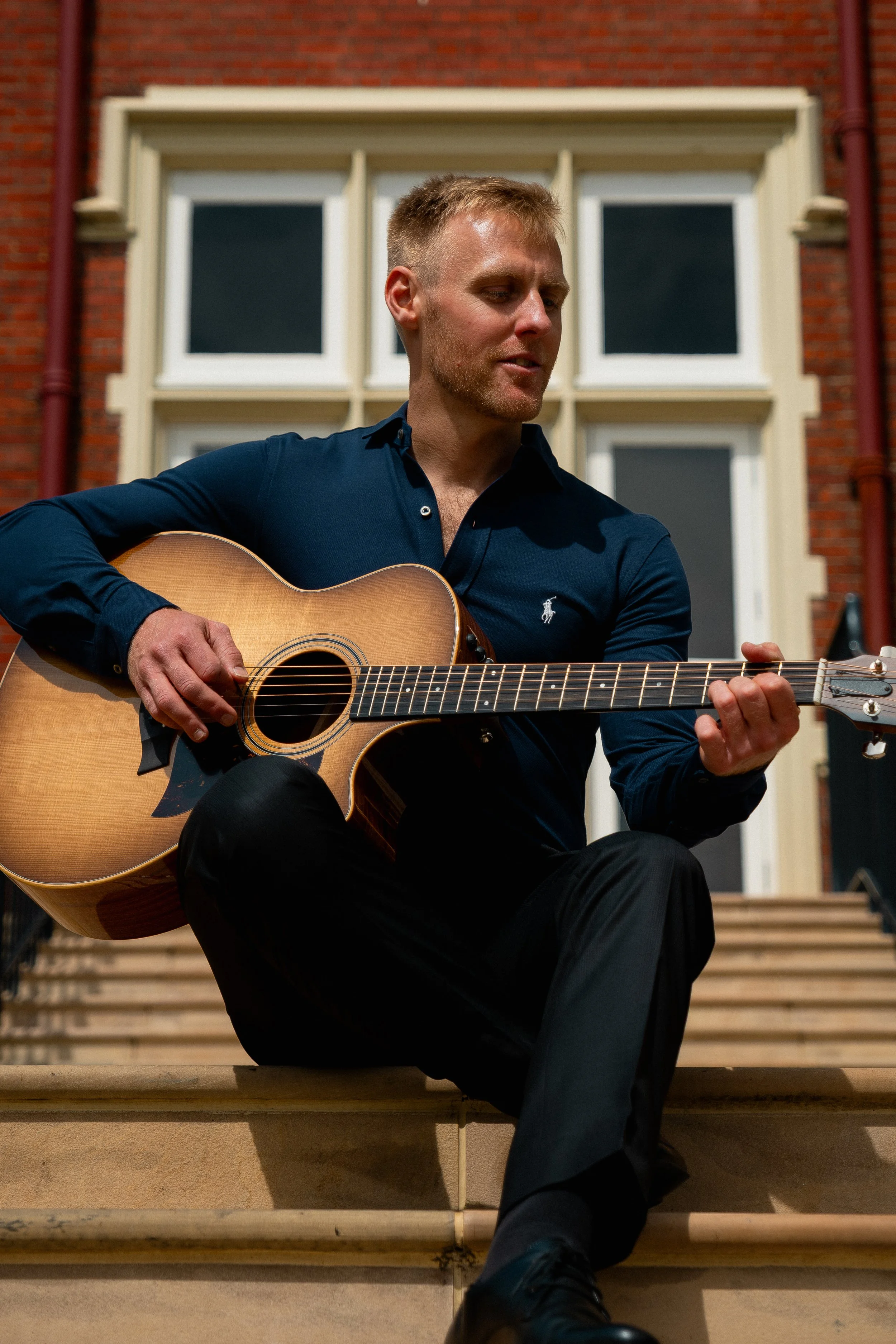 A man with short blonde hair sitting on outdoor steps playing an acoustic guitar in front of a building with large white window frames.