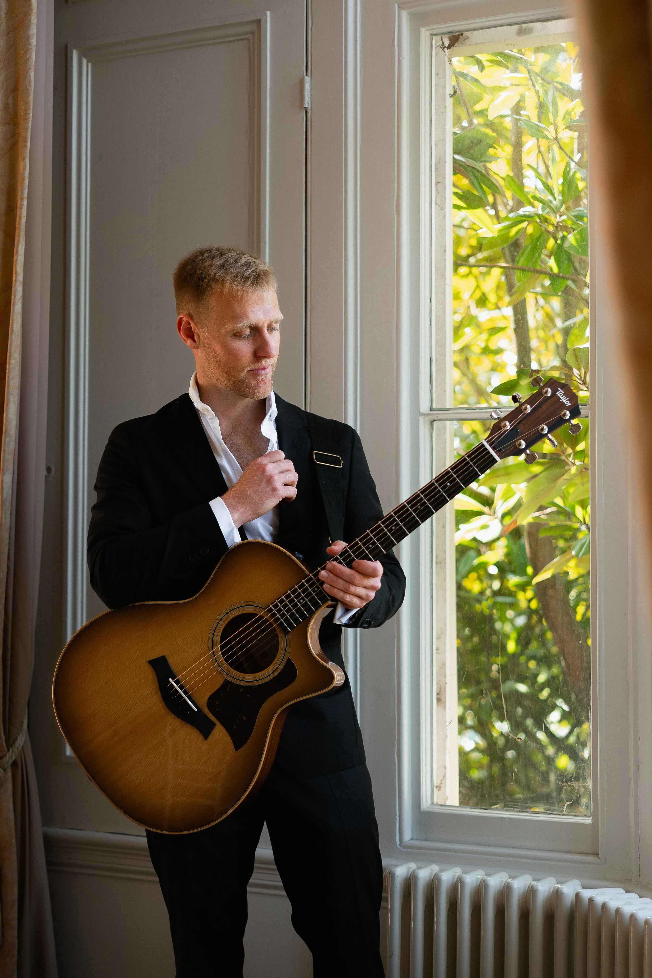 A man in a black suit playing an acoustic guitar on a windowsill inside a room, with greenery outside the window.