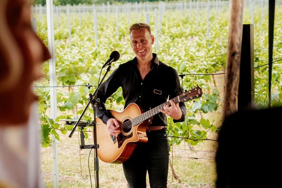 A musician strumming a guitar at a wedding surrounded by rows of grapevines at a sunny vineyard venue.