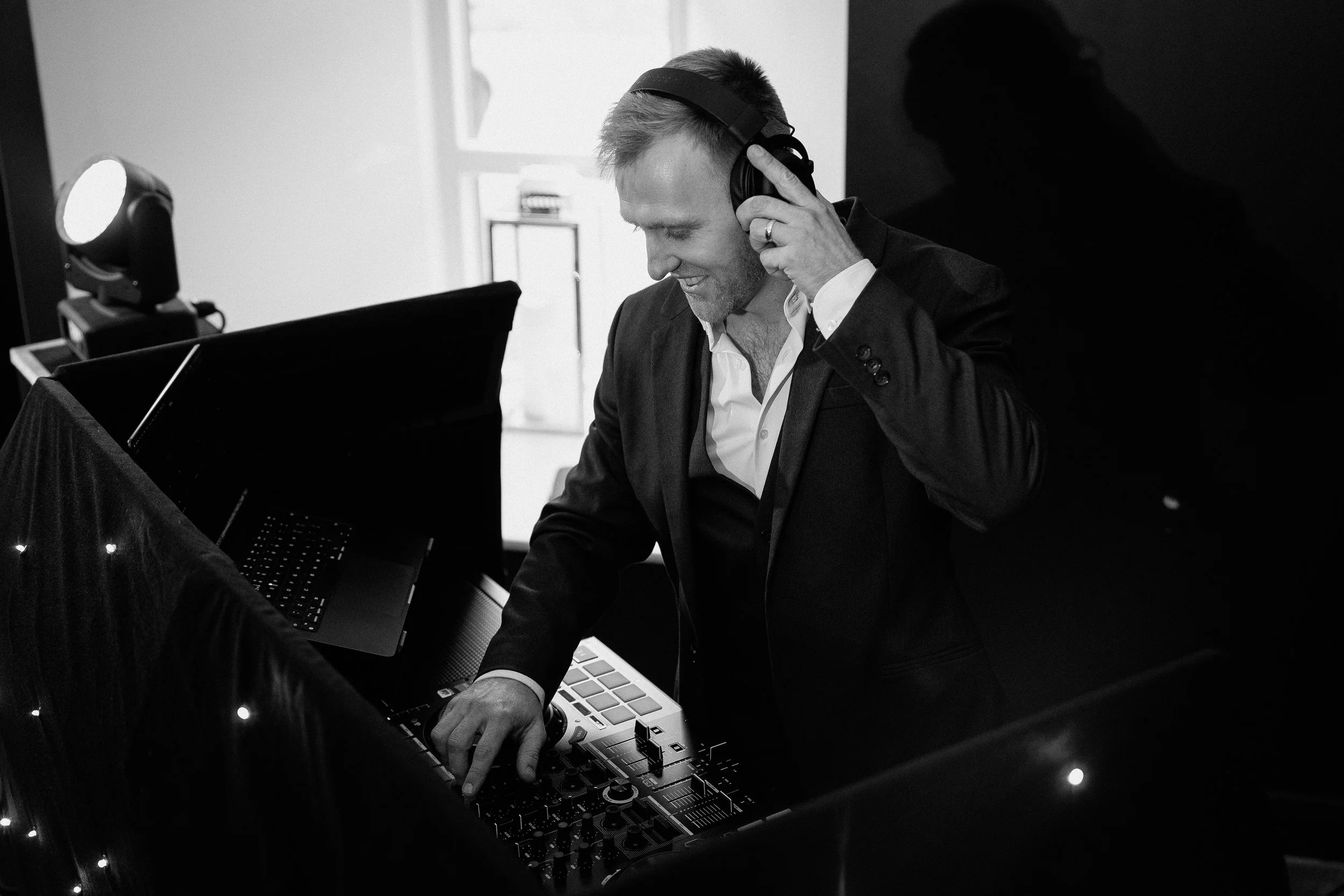 A man in a suit and white shirt wearing headphones and smiling as he mixes music on DJ equipment in a well-lit room.