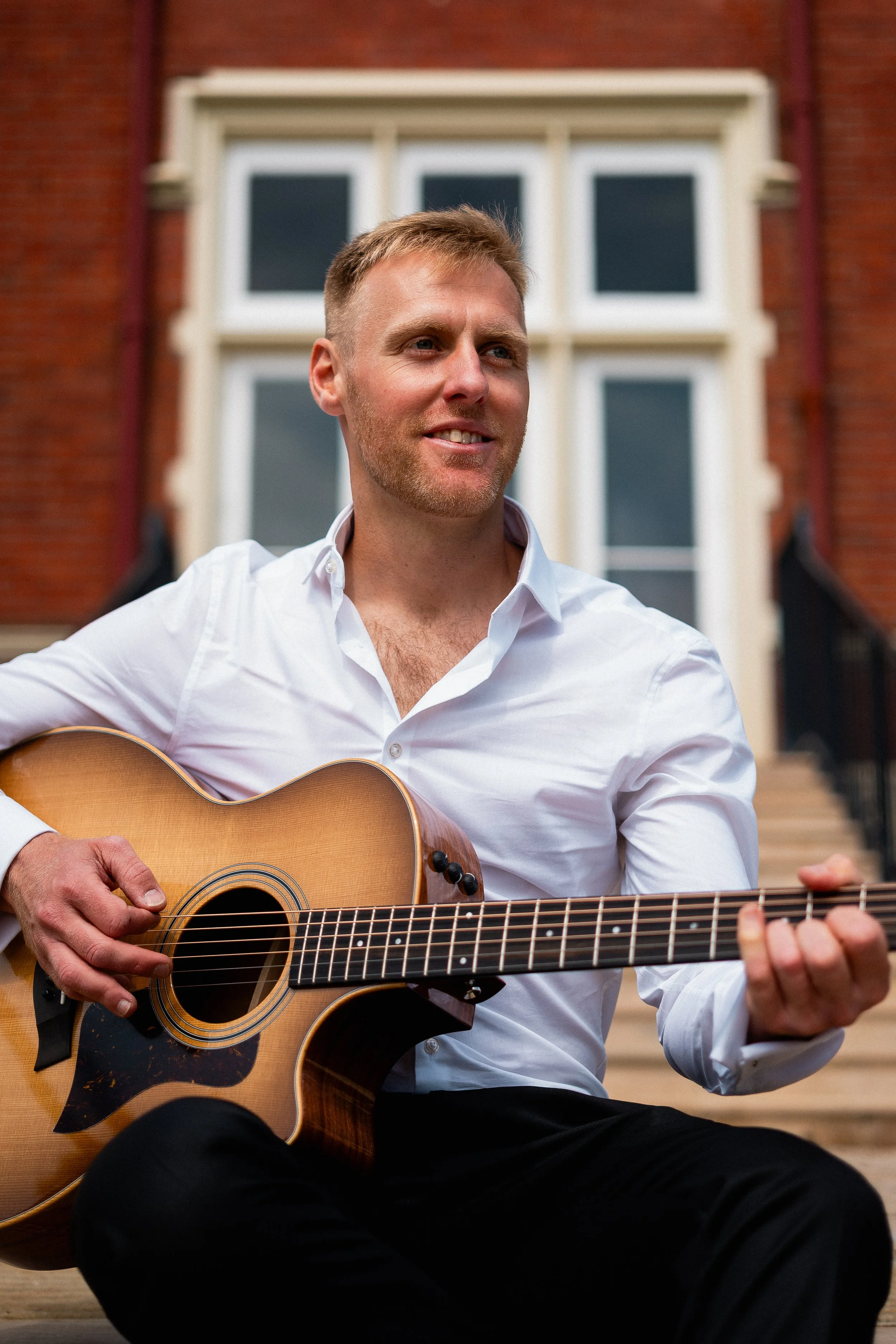 A man in a white shirt playing an acoustic guitar while sitting on steps outside a brick building.