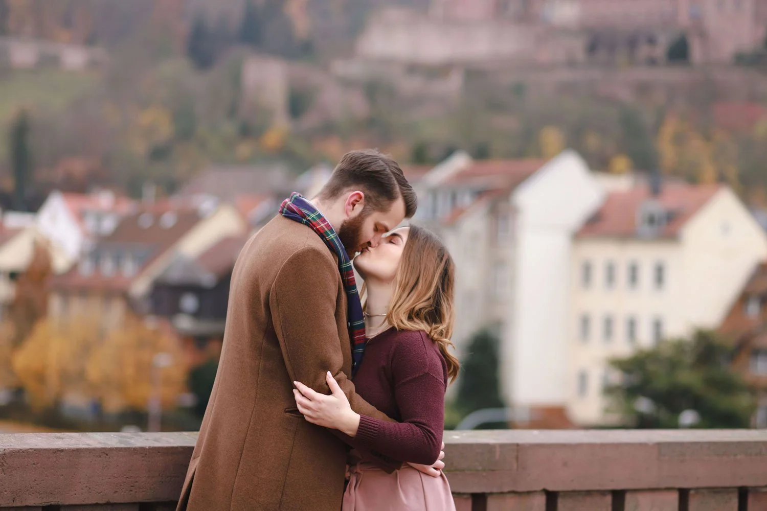 A couple kisses on an old bridge in Heidelberg with a cityscape in the background.