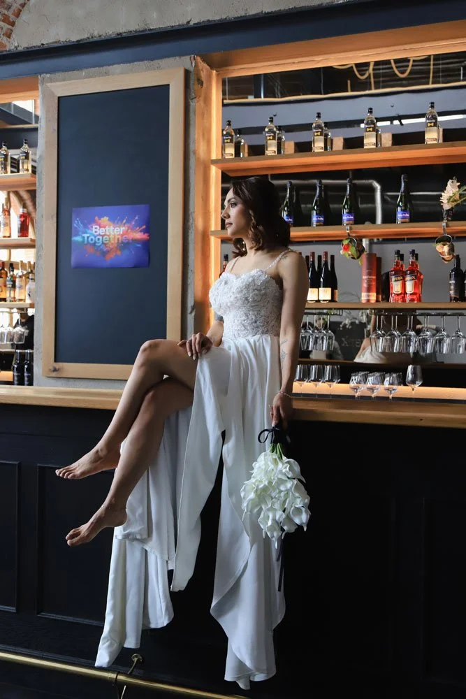 Eine Braut trägt ein weißes Kleid und sitzt fröhlich auf der Bar, umgeben von festlicher Stimmung.