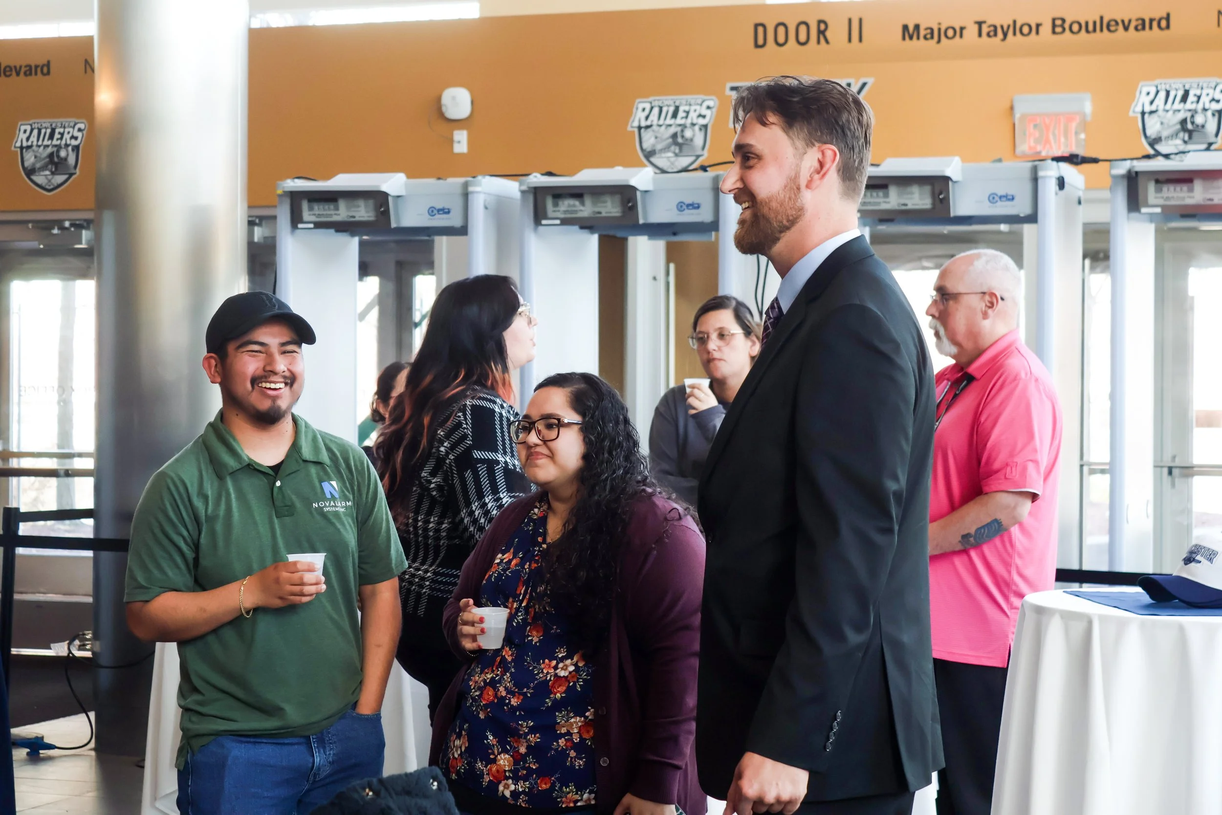 Group of people engaging in conversation at an indoor event near a table. A smiling man holding a paper cup, a woman with glasses and dark curly hair, a man in a suit with a beard, and others in the background. There are signs with logos and text in 