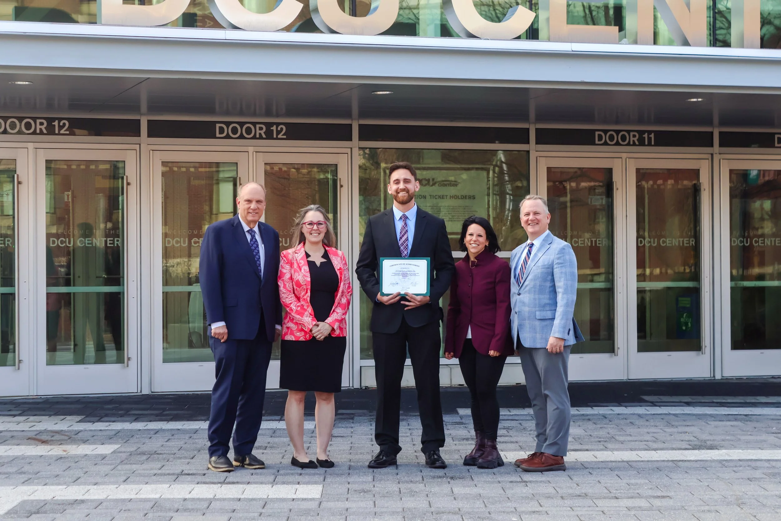 Group of five people standing in front of a building with glass doors and signs indicating doors 11 and 12. One person in the middle is holding a framed certificate. All are dressed in business attire, smiling and posing for the photo.