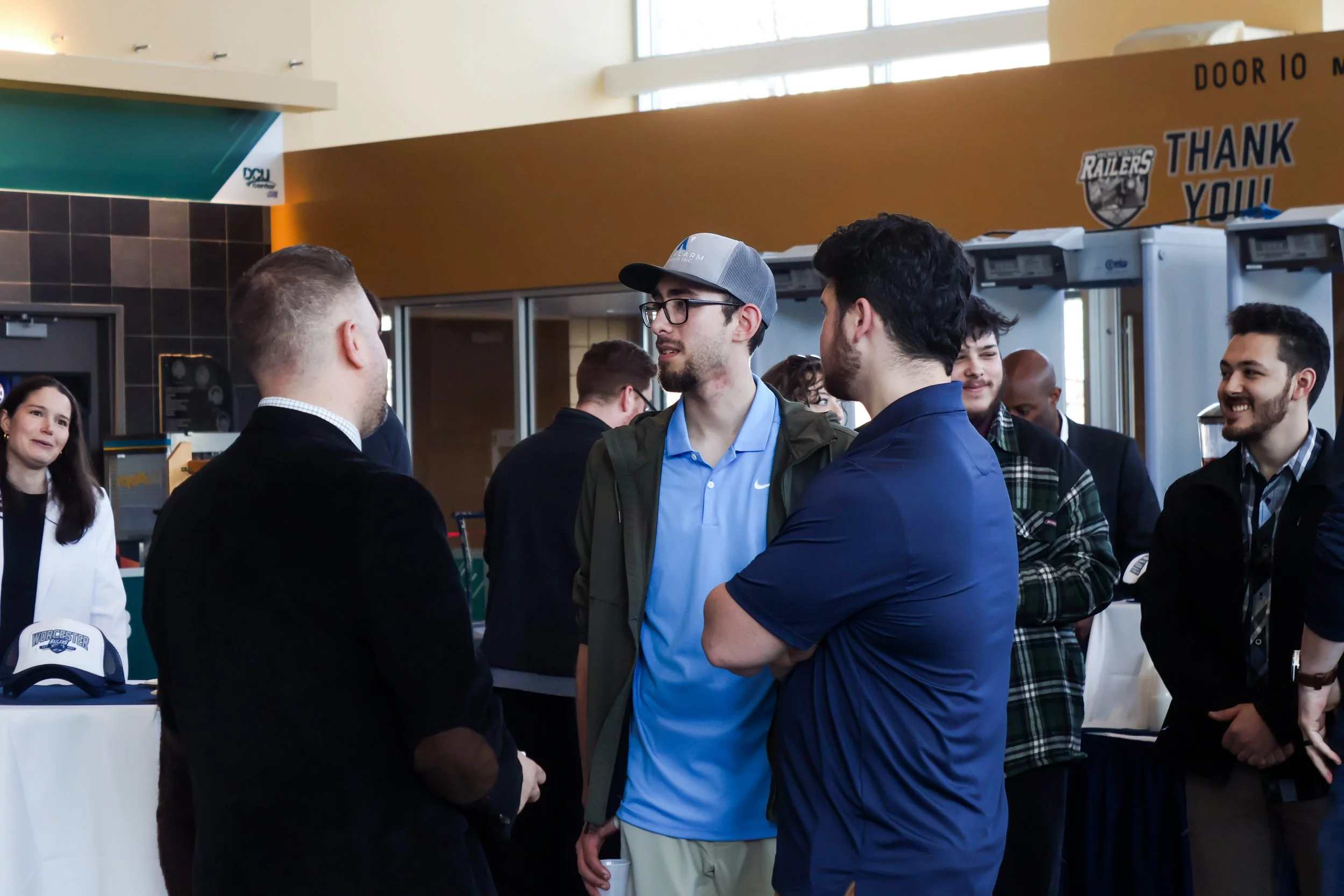 Group of people talking and socializing at an indoor event or conference, with some wearing sports caps and branded apparel.