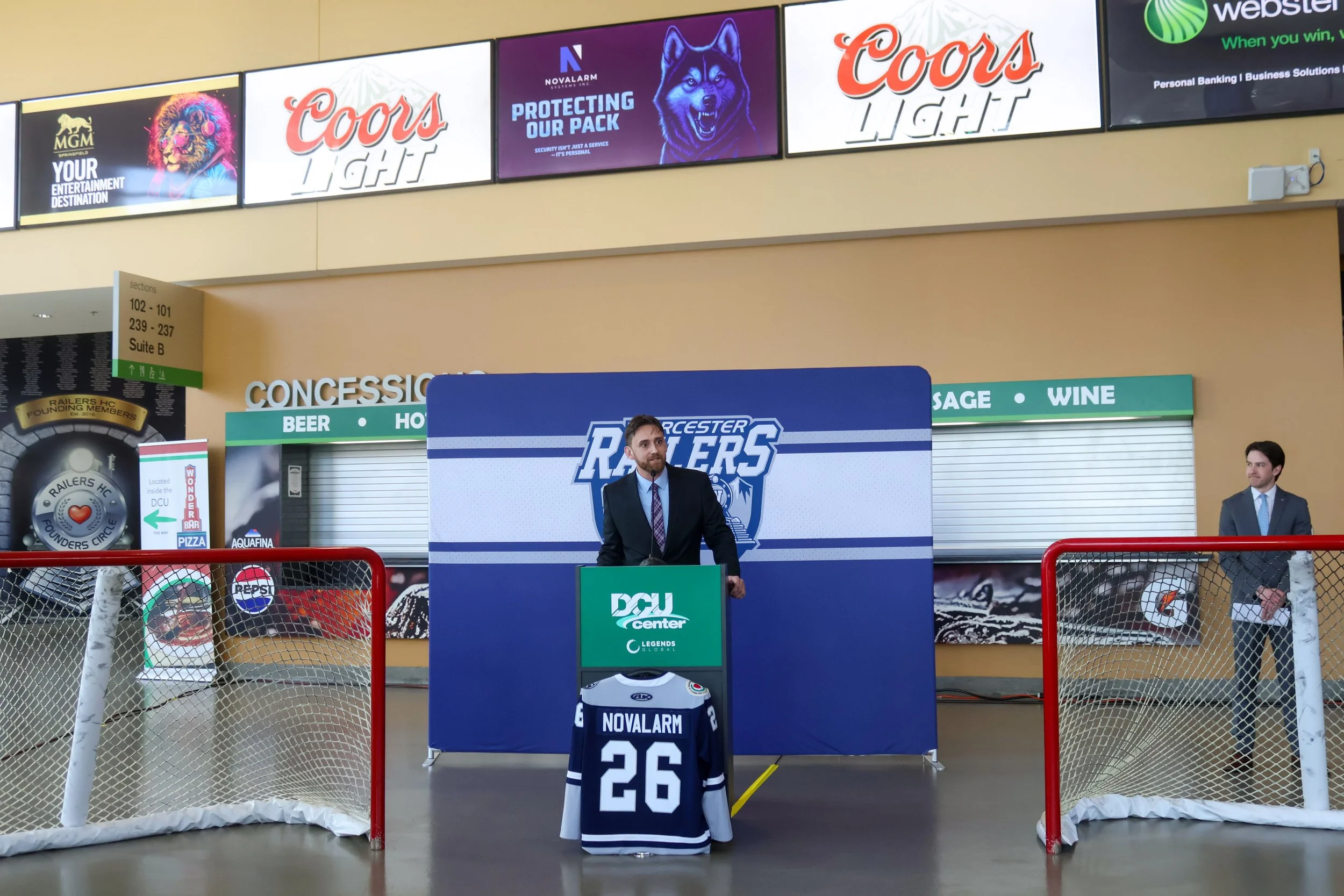 An indoor hockey arena with promotional displays and digital screens above. In the foreground, a hockey jersey with the name "Novalarm" and the number 26 is on a stand. Two men in suits are standing behind a table as part of a presentation or event.