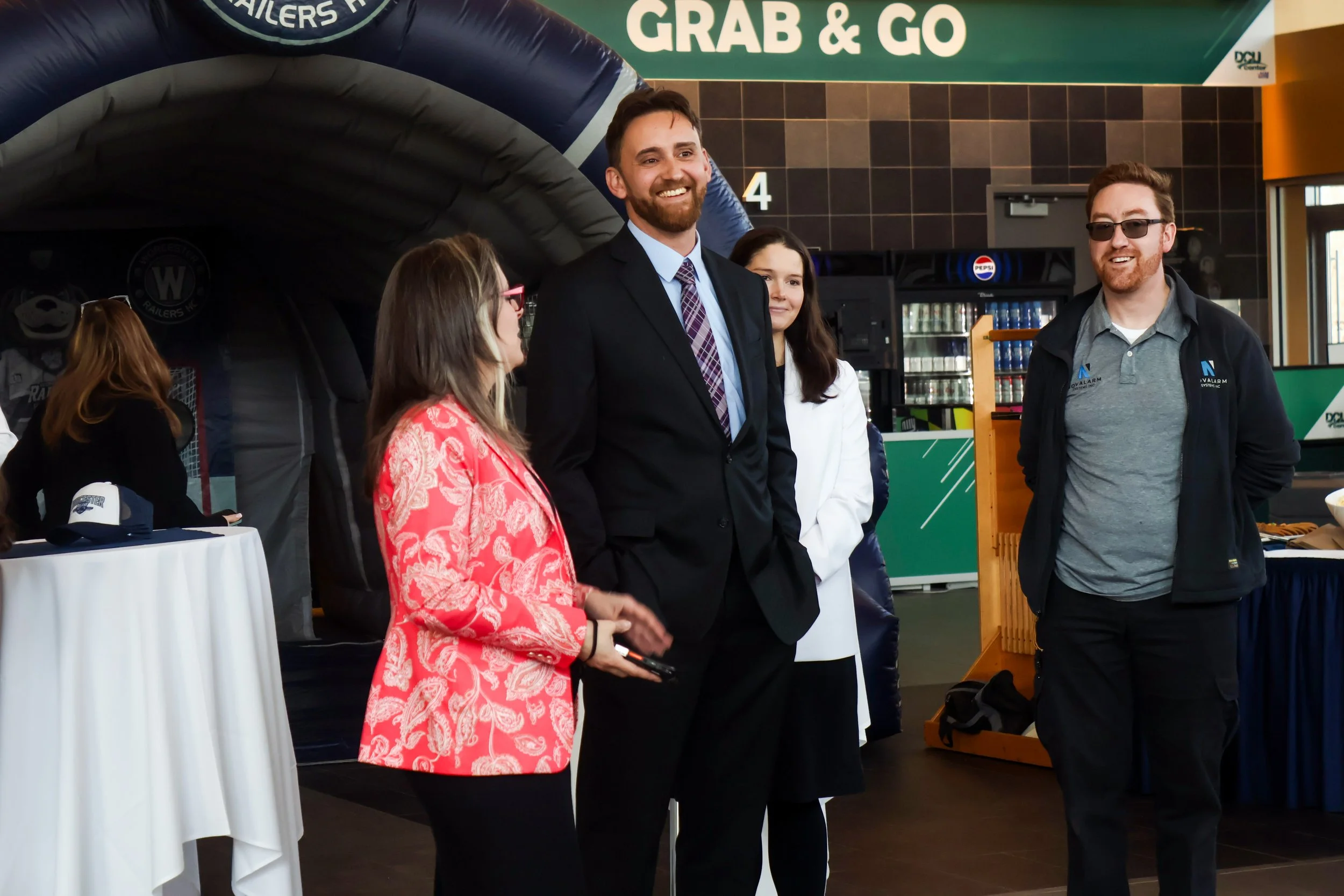 Group of people at a promotional event, including a woman in a red patterned dress talking to a bearded man in a dark suit, and a man with glasses and a beard wearing a gray polo shirt, with a green sign reading 'GRAB & GO' in the background.