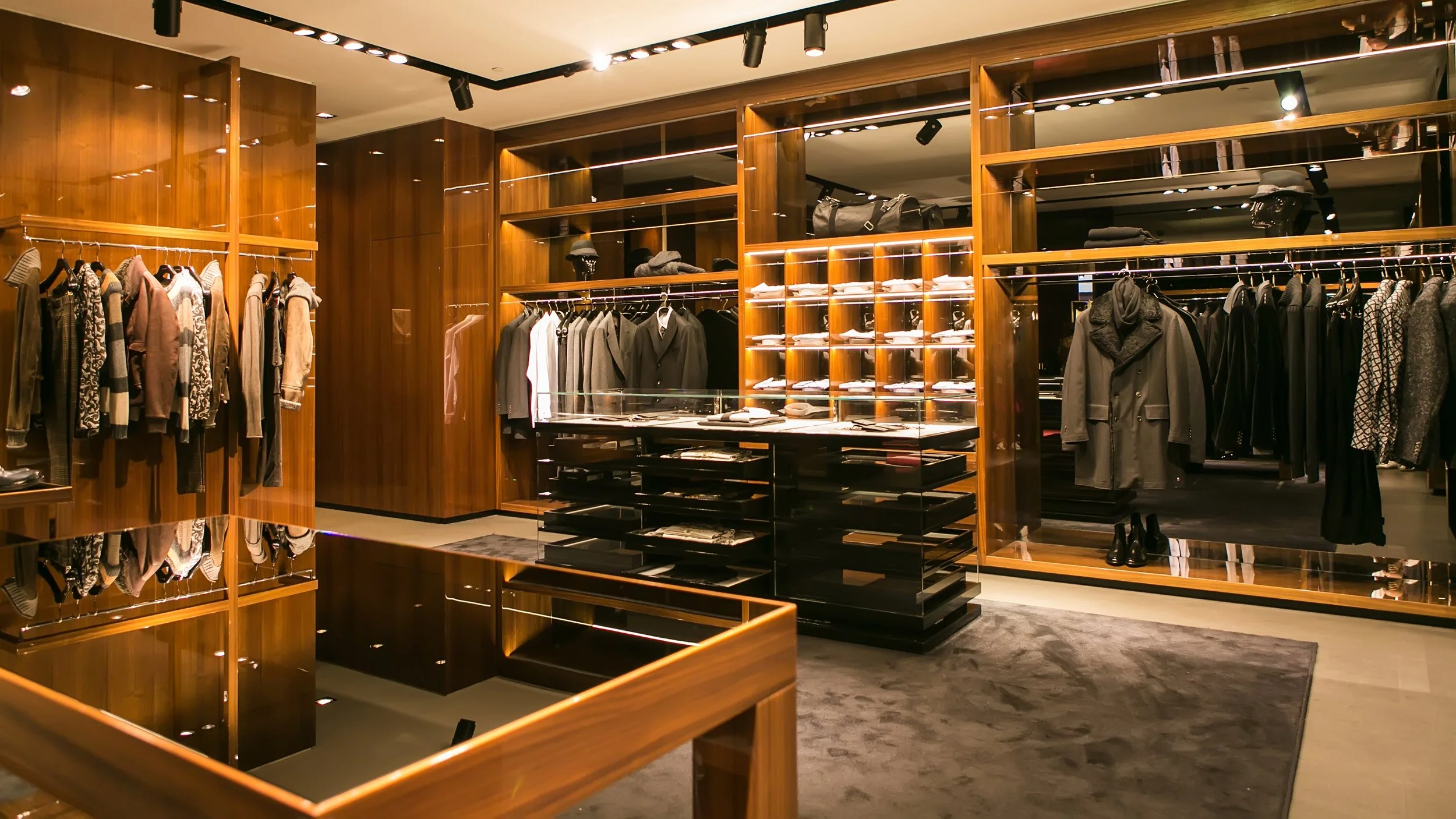 Interior of a luxury clothing store with wooden shelves and clothing racks displaying various men's apparel including suits, jackets, and shirts.
