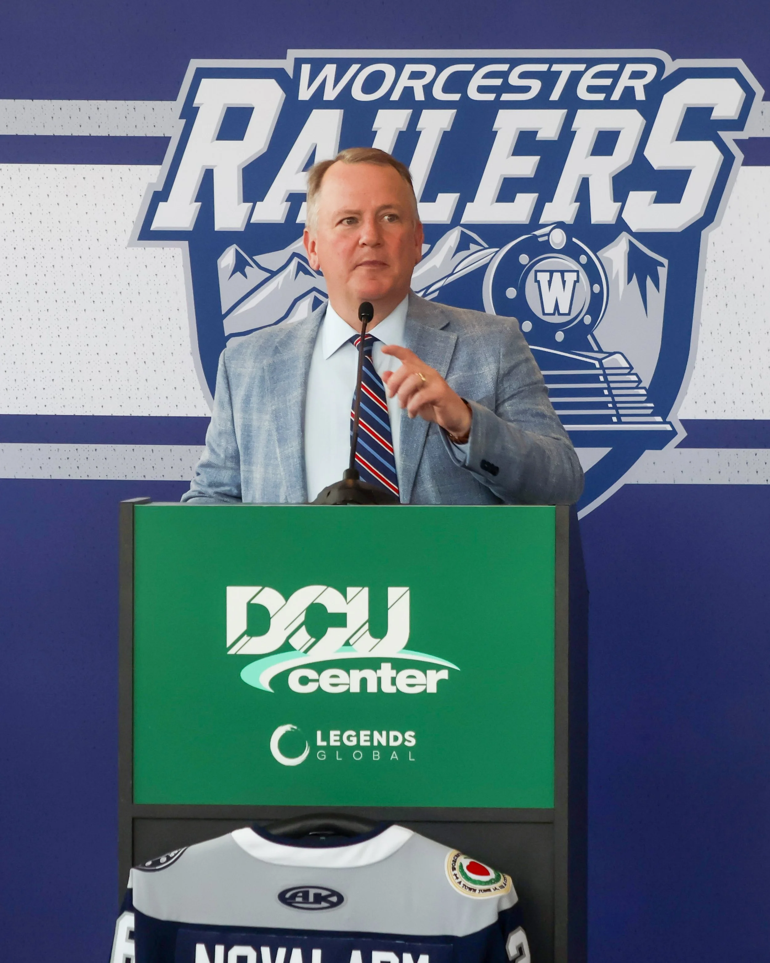 An individual standing at a podium with a microphone, in front of signage for Worcester Railers and DCU Center, wearing a light grey suit and striped tie. The background features logos and text for Legends Global and D.C. United.