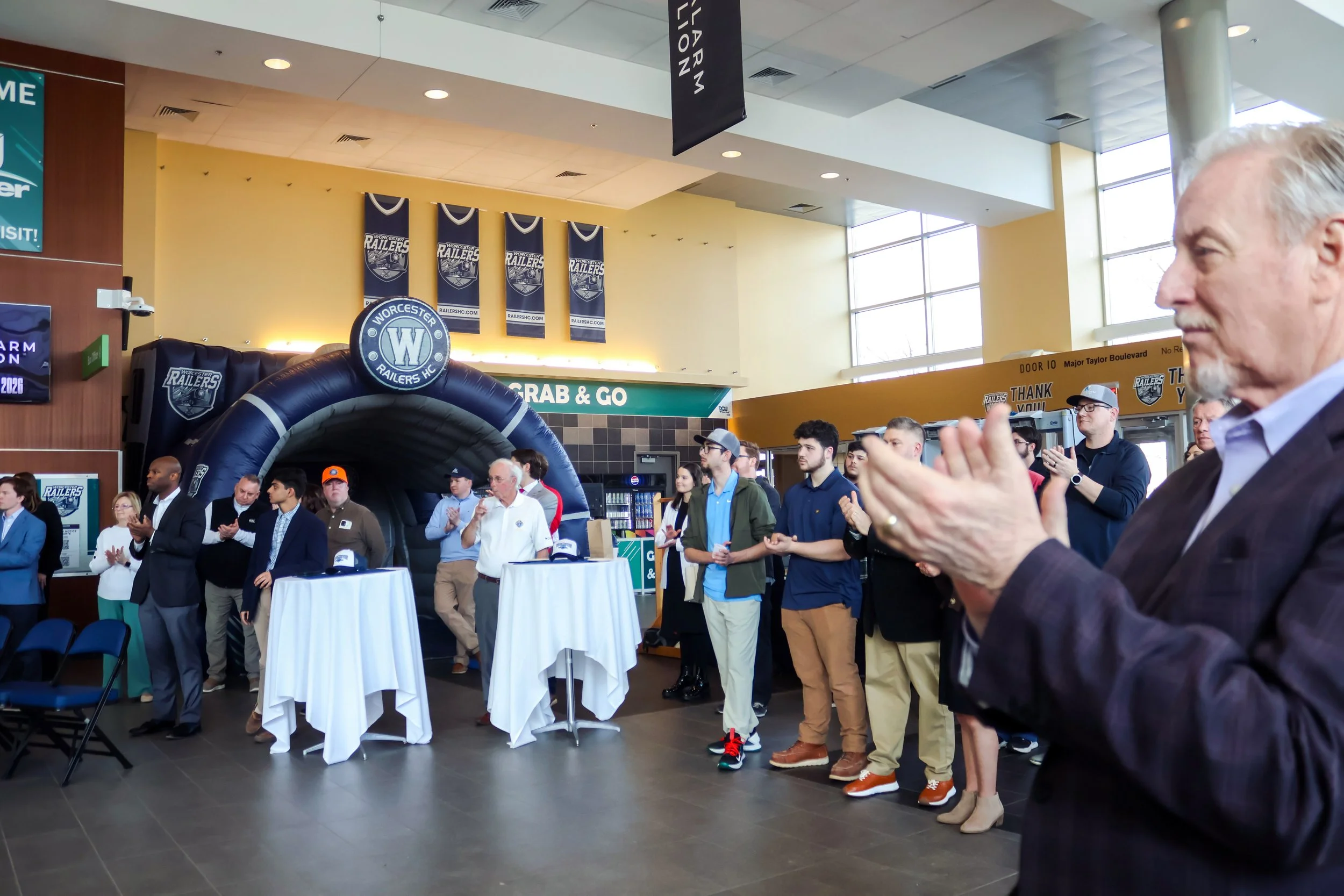 Group of people at a hockey event inside a building with banners and signs for Worcester Railers, some are applauding or listening attentively, with tables and chairs nearby.