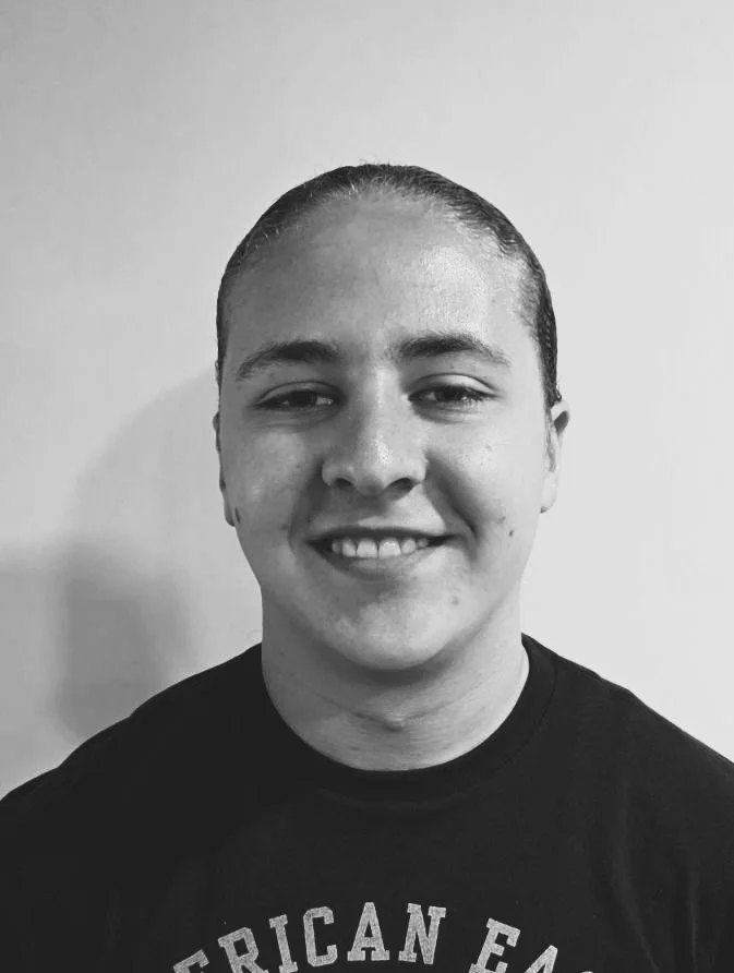 A black-and-white portrait of a young man with slicked-back hair, smiling, wearing a dark T-shirt in front of a plain wall.