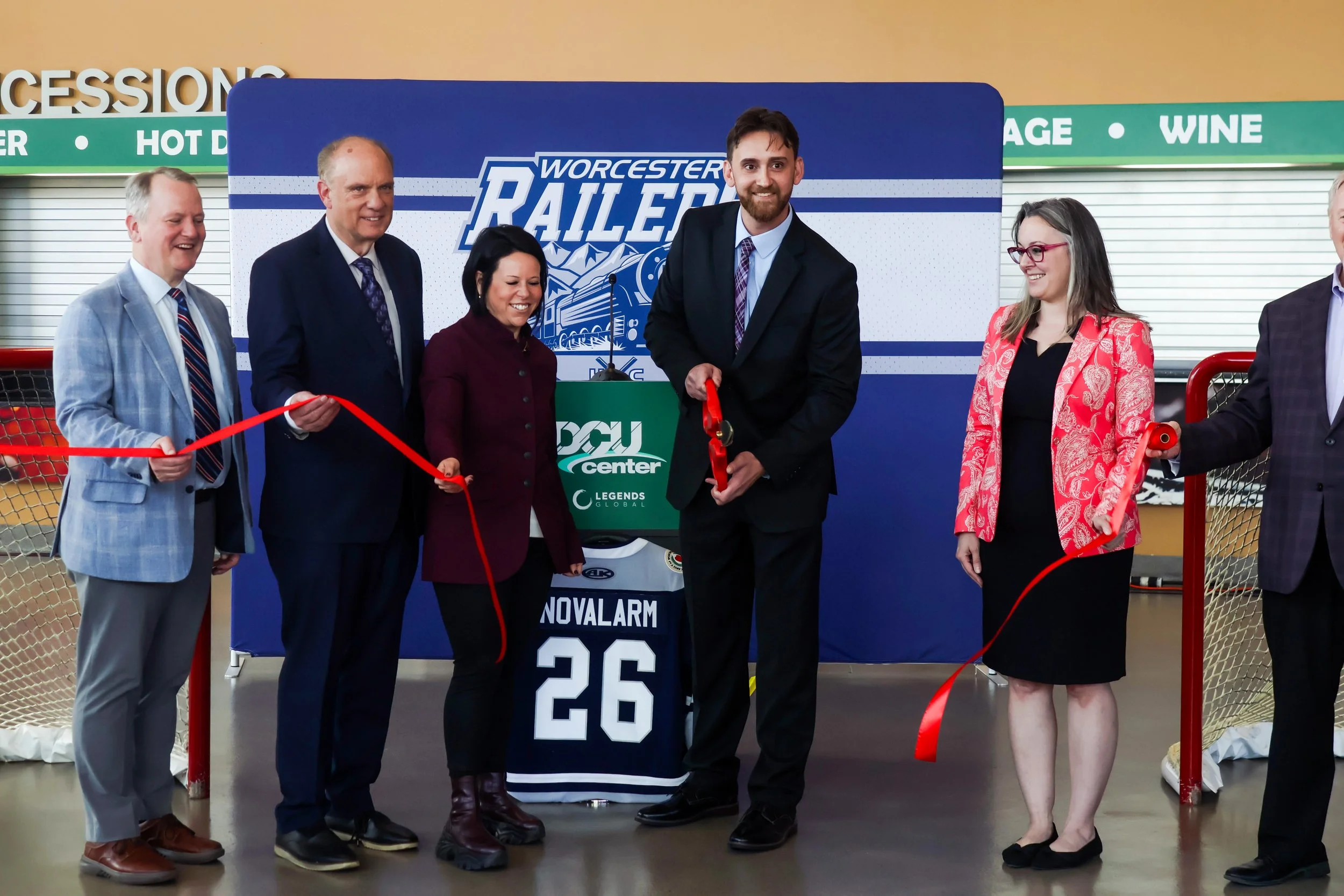 Group of six people standing at a ribbon-cutting ceremony inside an arena, with a backdrop displaying 'Worcester Railers'.