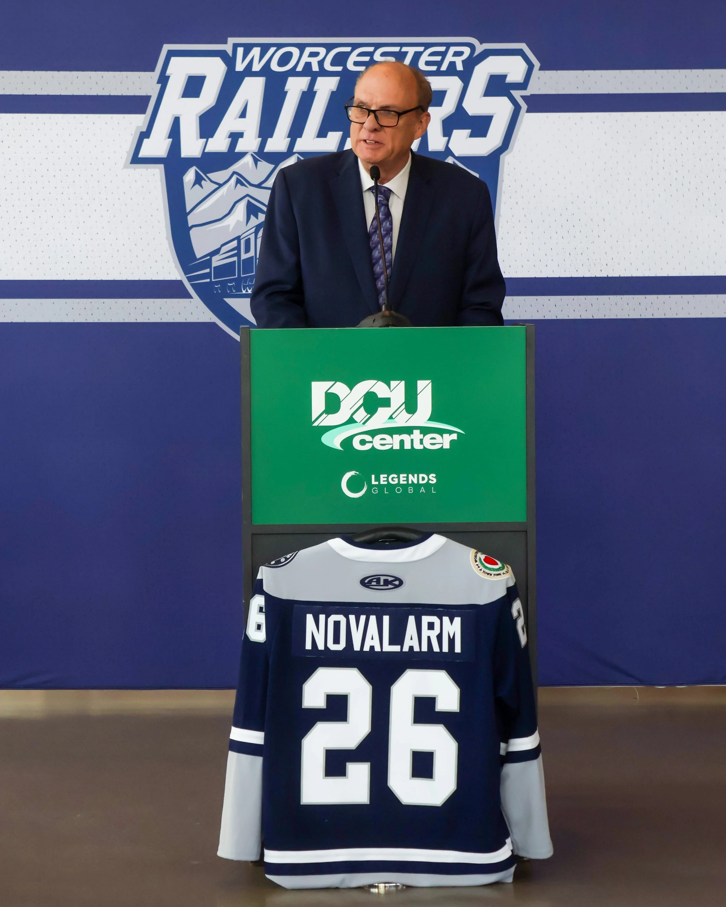 Press conference at Worcester Railers hockey rink with a man in a suit speaking at a podium, with hockey jersey on display in front of the podium. The background features the Worcester Railers logo and a mountain scene.