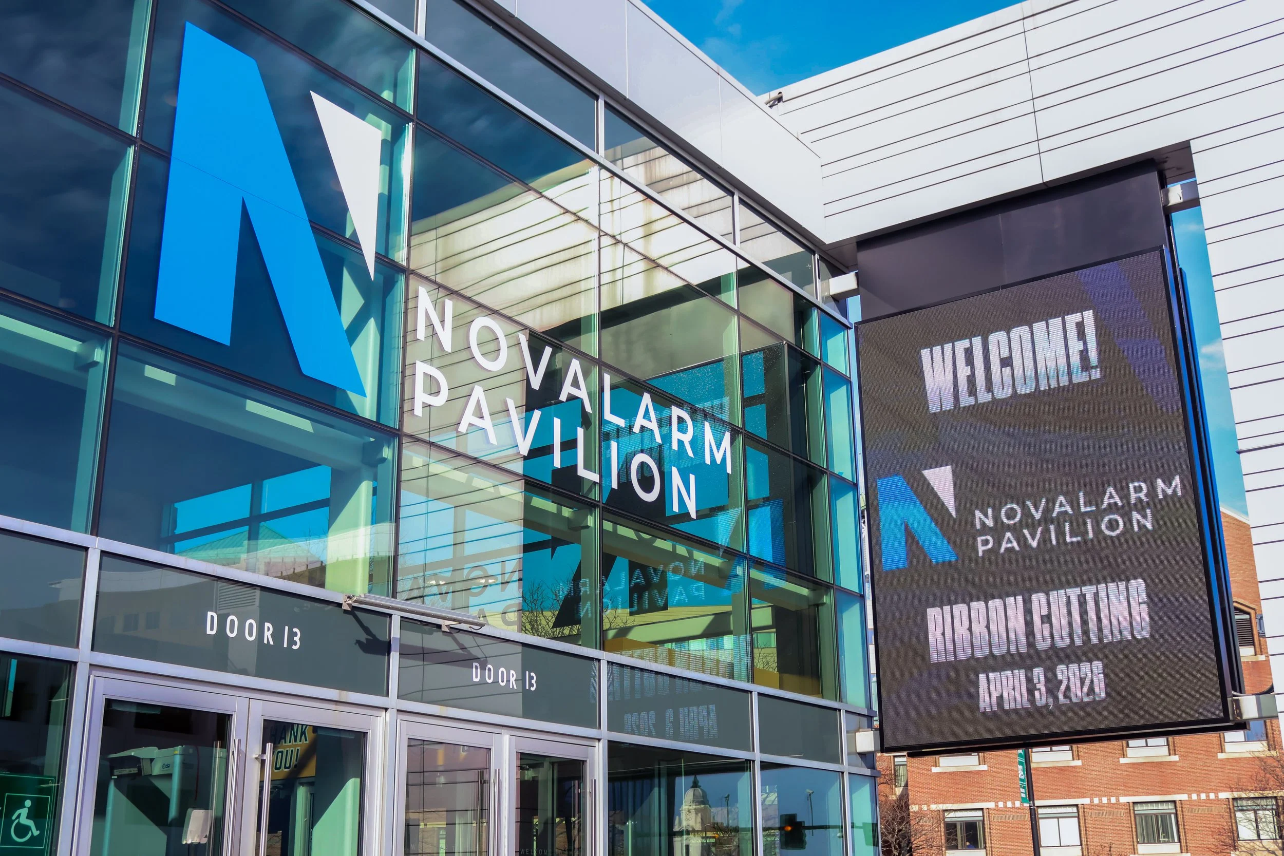 Exterior of Novalarm Pavillon building with large glass windows, a digital welcome sign, and signs for doors 13 and 14. Clear blue sky and part of another building in the background.